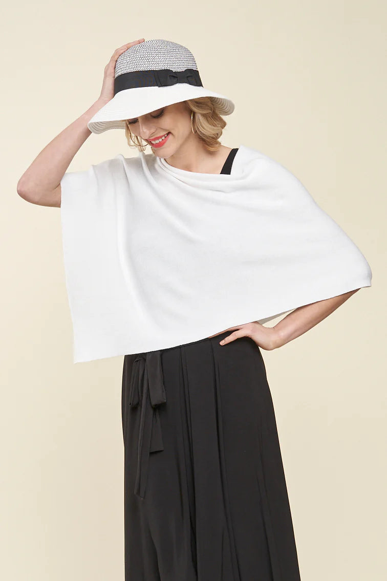 A person stands against a plain background wearing Parkhurst’s Capri Poncho, styled as a white asymmetric top, paired with black pants and a wide-brimmed hat.