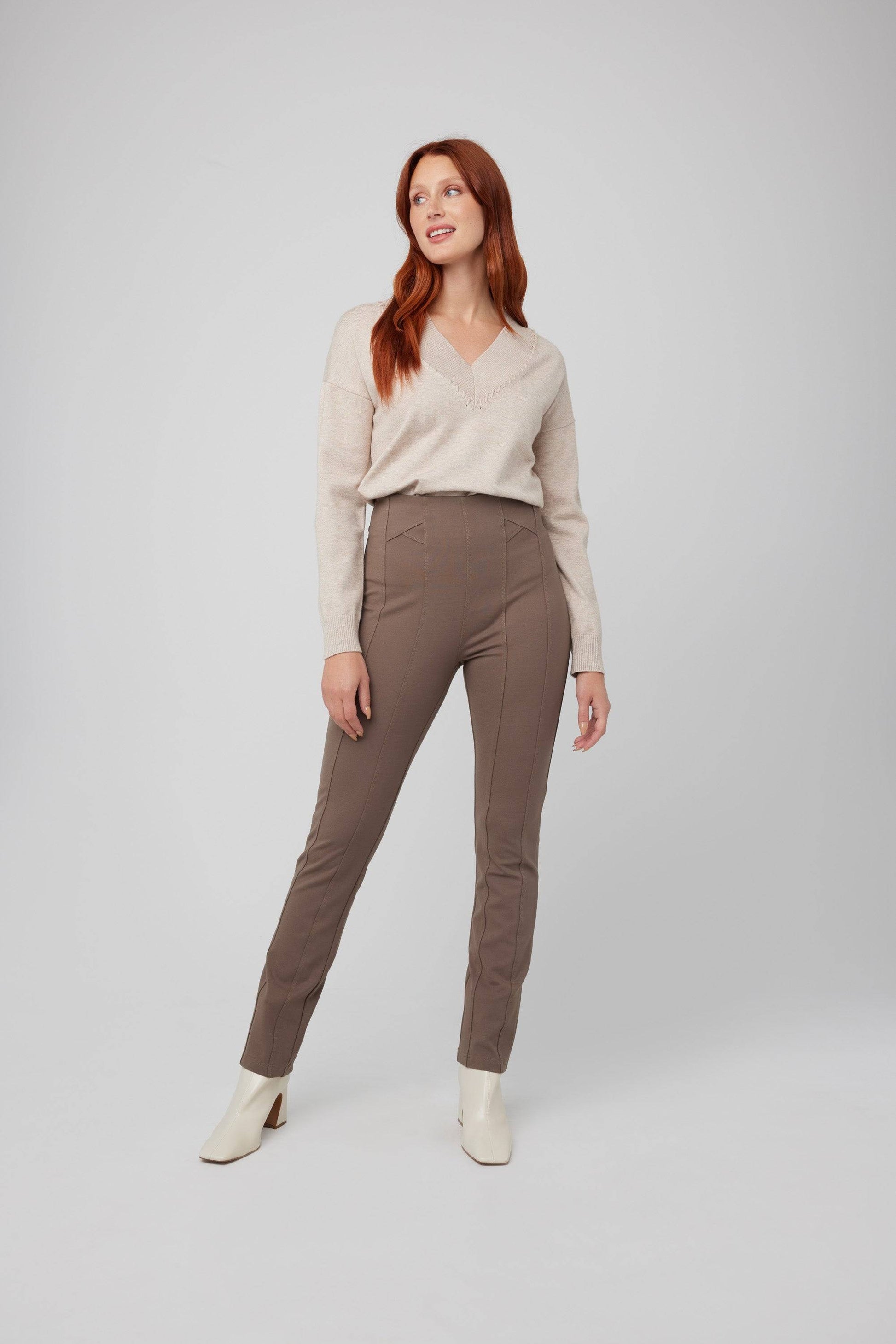 A person with long red hair is standing and looking to the side, wearing a light beige long-sleeve top, brown high-waisted Front Seam Slim Pull On Pant by CoCo Y Club, and white ankle boots. The background is gray.