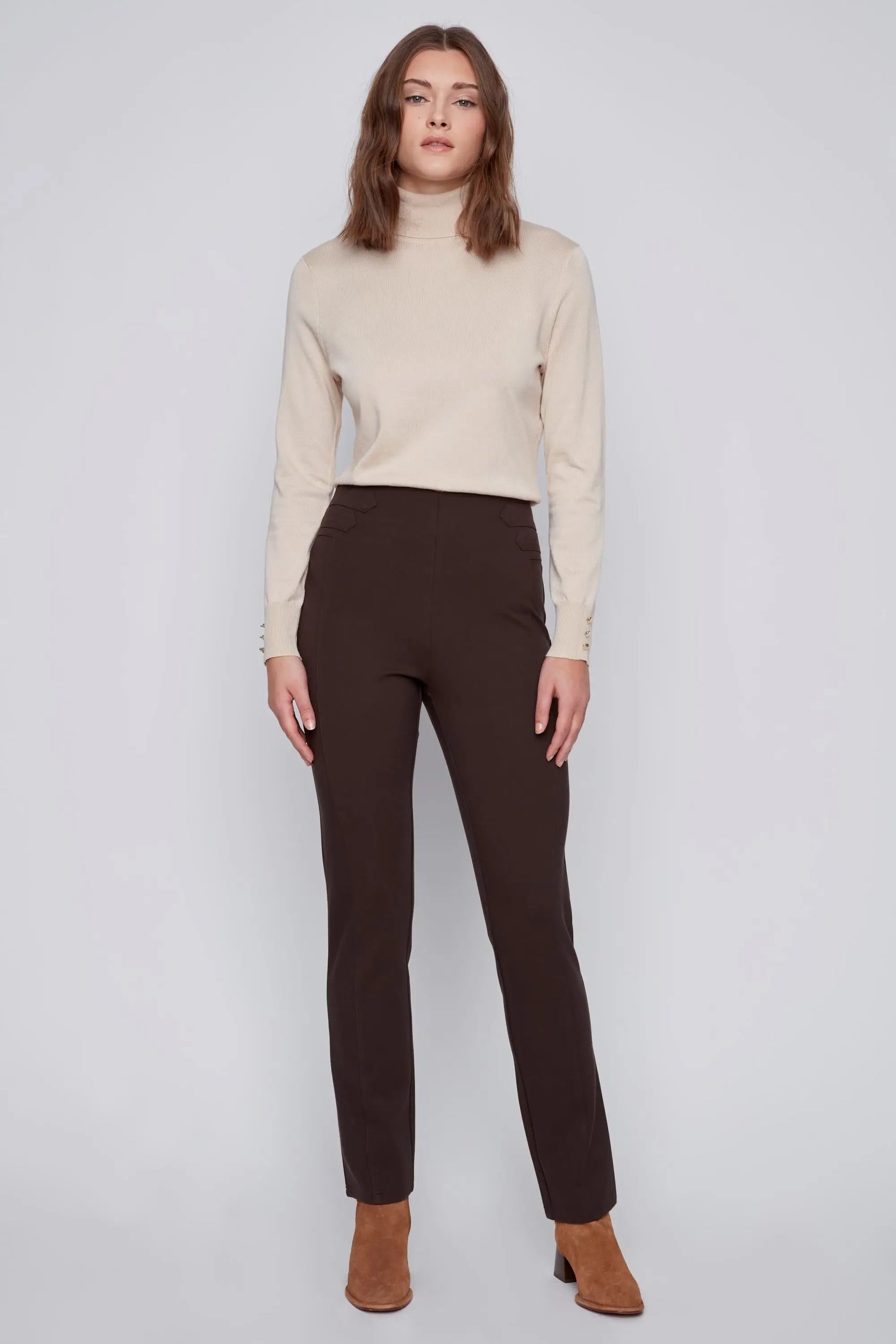 A person with long hair stands against a plain background, showcasing effortless style in a beige turtleneck sweater, dark brown high-waisted CoCo Y Club Pull On Pant, and brown ankle boots.