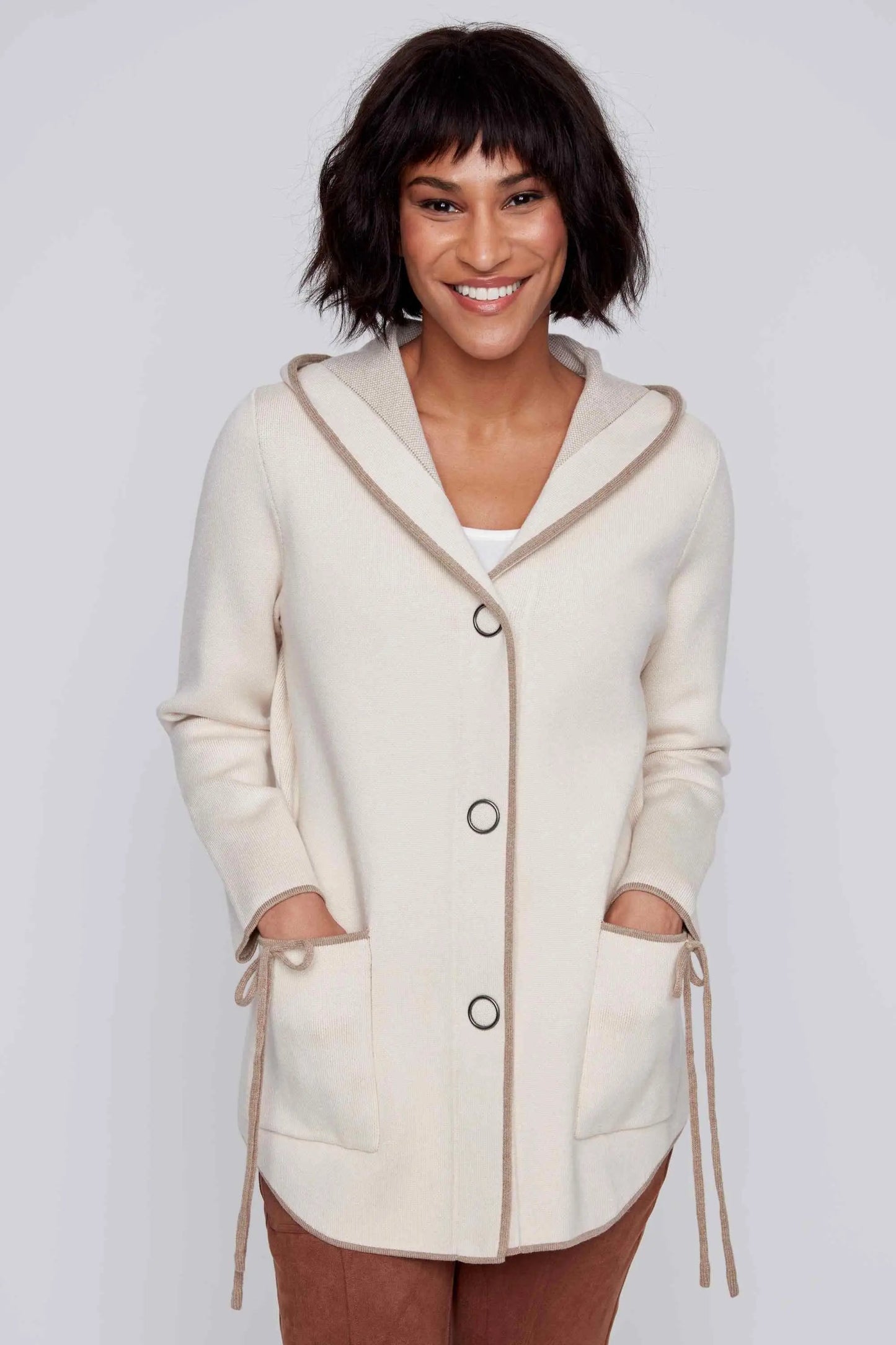 A woman smiles while wearing the Snap Front Hooded Knit Jacket from CoCo Y Club, which is cream-colored with brown accents, front buttons, and pockets. She has short, dark hair and a white top underneath.