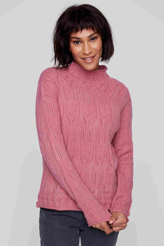 A person with short dark hair wears a trendy pink High Neck Cable Knit Sweater from CoCo Y Club made of super soft material, paired with dark pants, standing against a plain gray background and smiling.