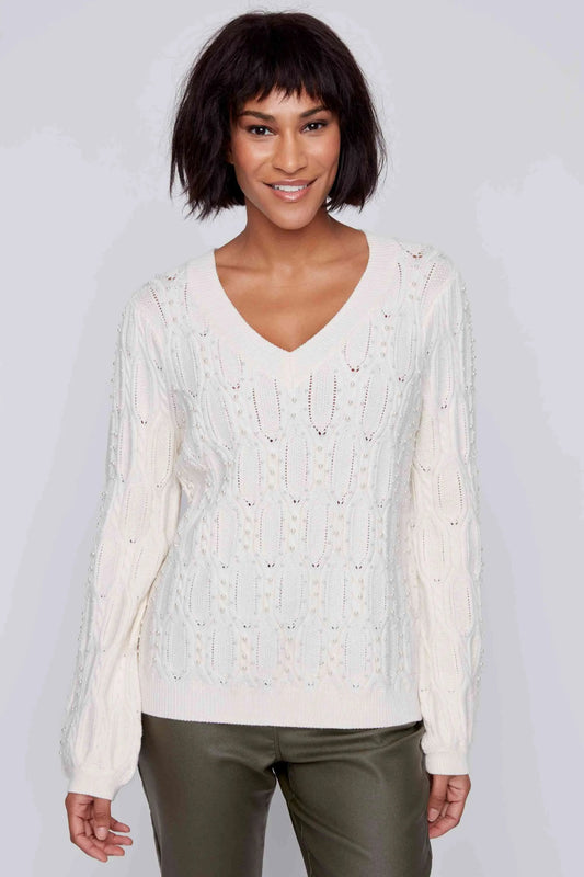 A person with short hair is wearing a CoCo Y Club Beaded Ivory Sweater featuring an intricate V-neck design and green pants, standing and smiling against a plain background.