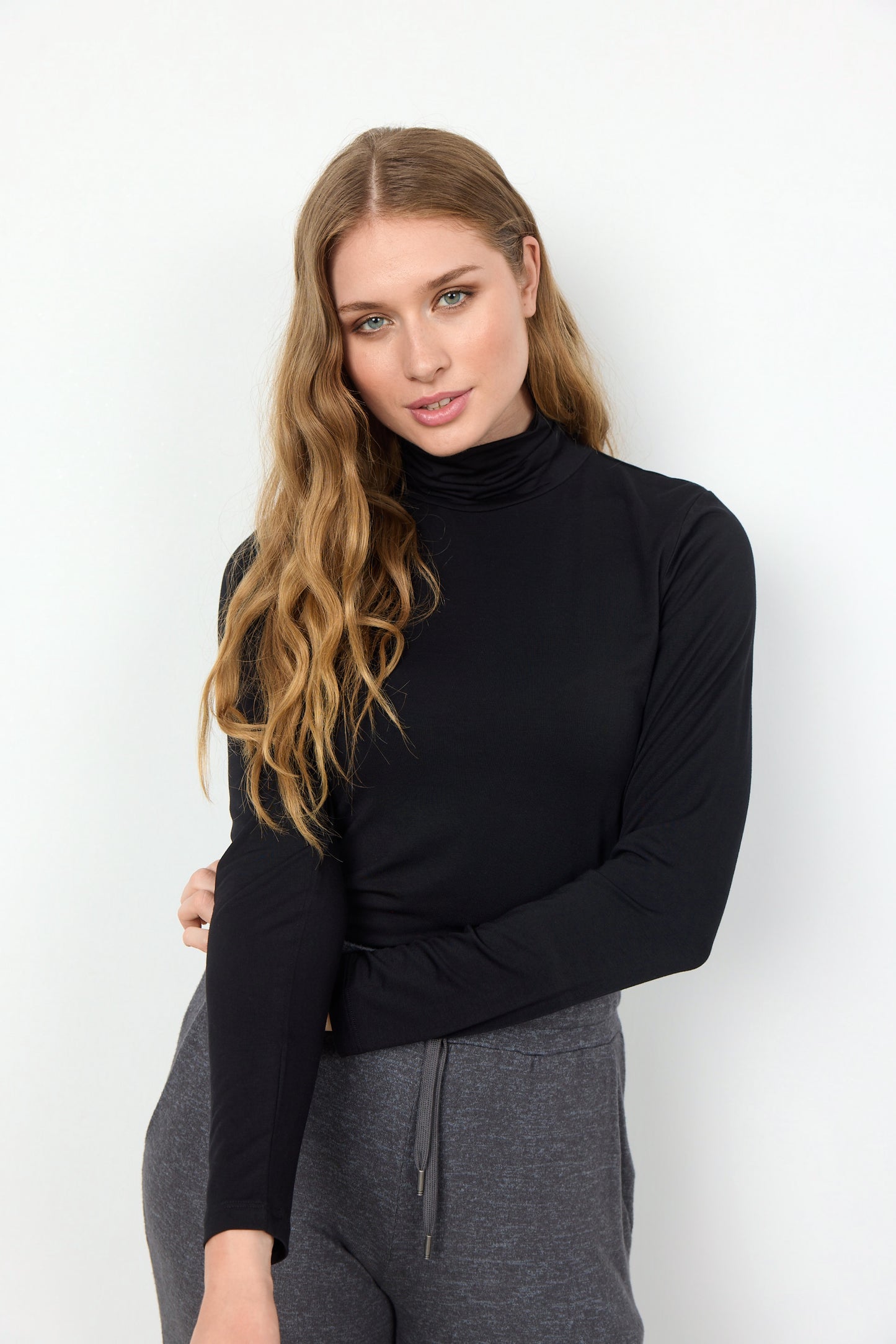 A stylish woman with long, wavy hair, dressed in a black Marica 12 Turtleneck from Soya Concepts and grey pants, stands against a plain white background.