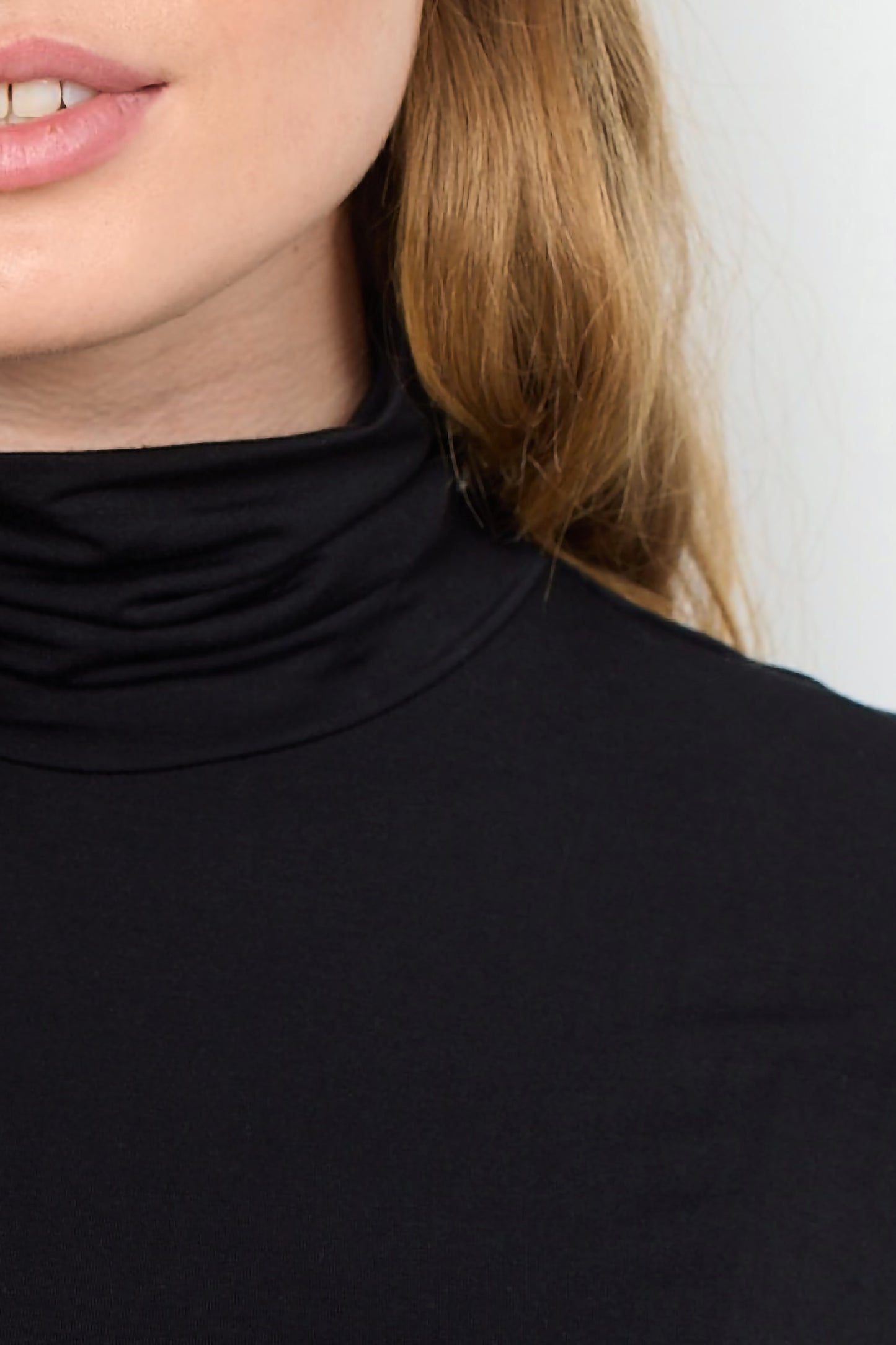 A stylish woman with long, wavy hair, dressed in a black Marica 12 Turtleneck from Soya Concepts and grey pants, stands against a plain white background.