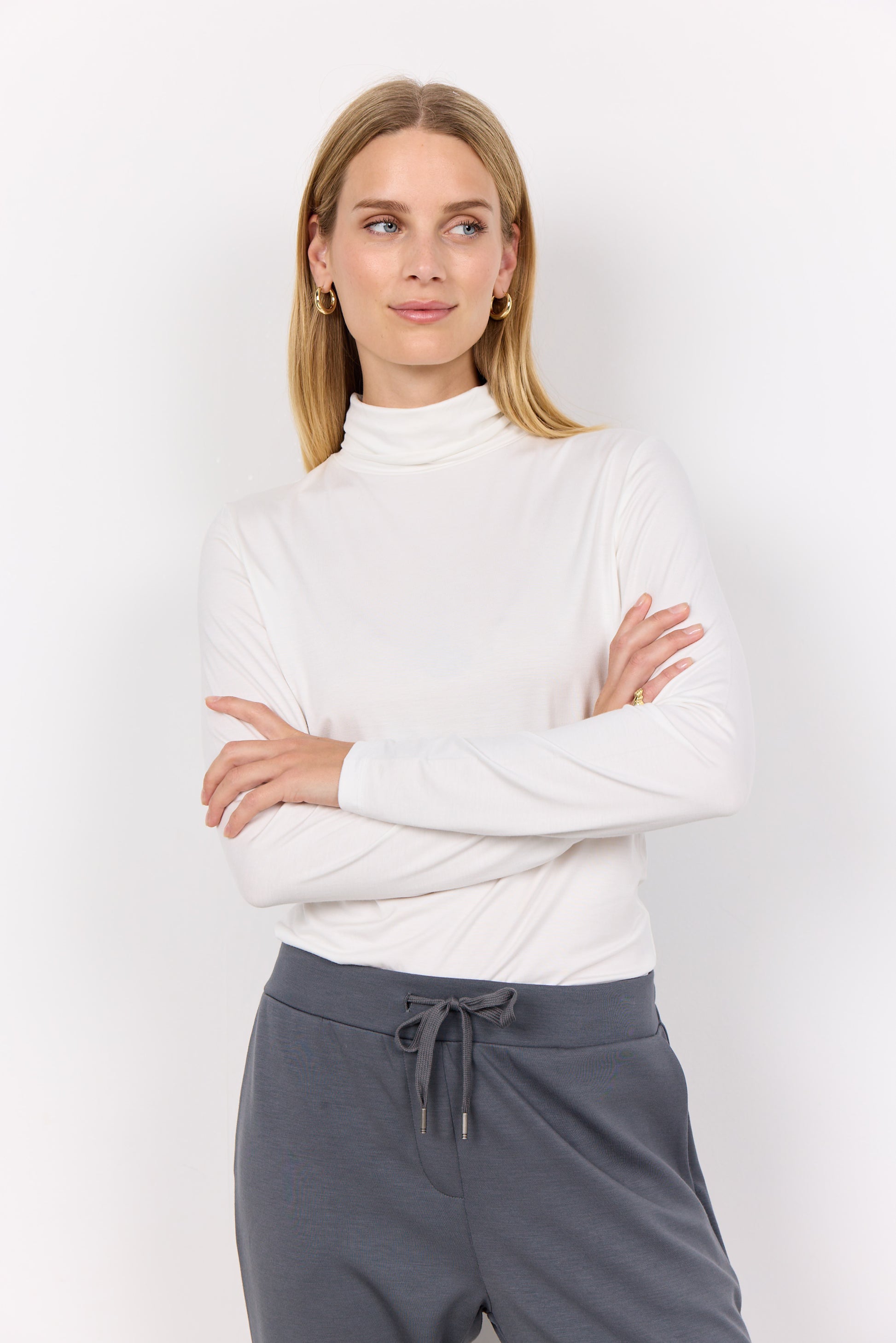 A stylish woman with long, wavy hair, dressed in a black Marica 12 Turtleneck from Soya Concepts and grey pants, stands against a plain white background.