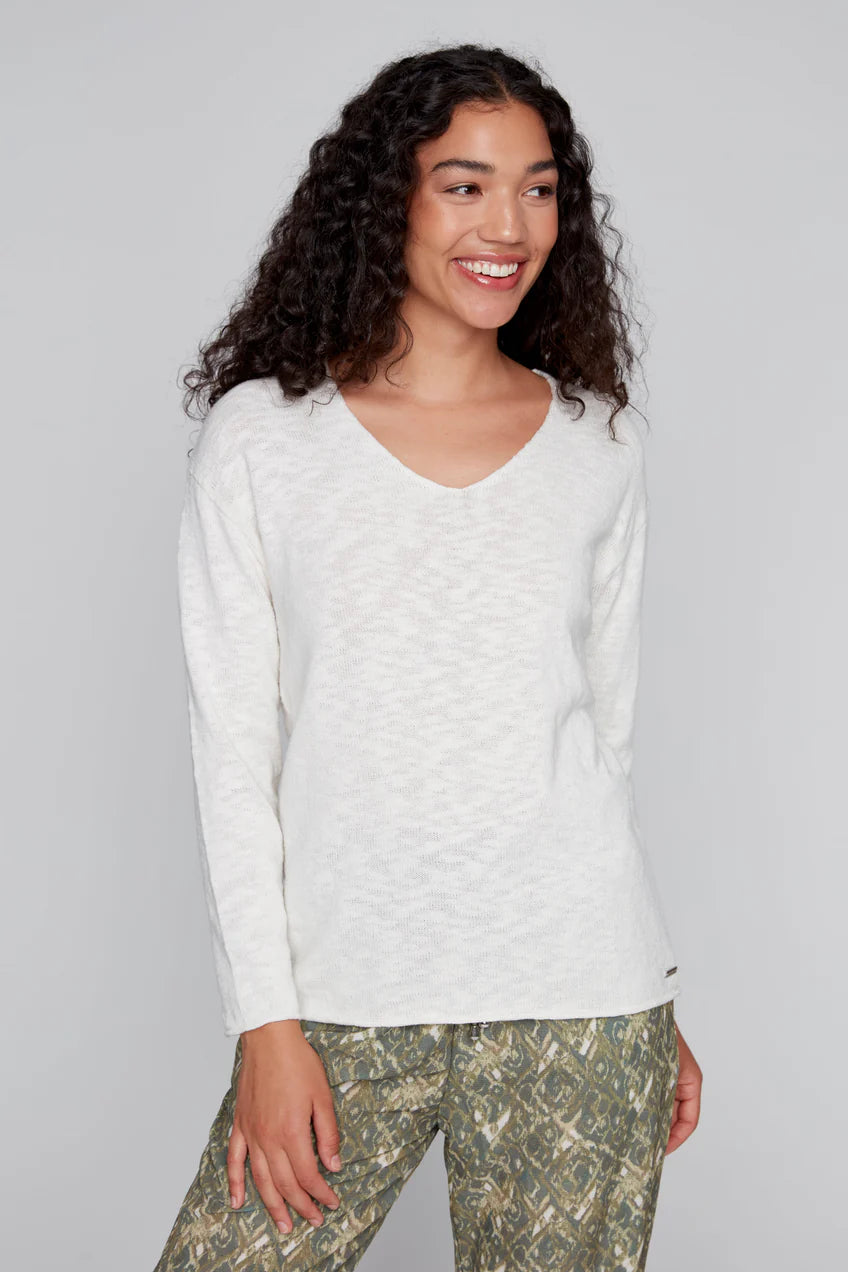 A smiling person with curly hair is dressed in an Off White Cotton Knit Sweater by CoCo Y Club and patterned green pants.