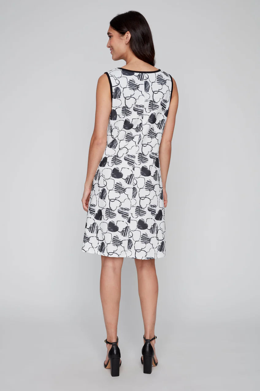 Against a gray background, the woman exudes confidence in her fashionable CoCo Y Club Black & White Heart Dress, characterized by its stylish two-tone design and sleeveless black and white floral pattern, paired elegantly with black heeled sandals.