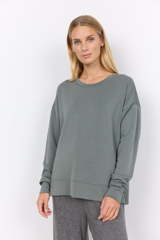 A person wearing a loose-fit Banu 164 Sweater by Soya Concepts in gray stands against a plain white background, paired with pants.