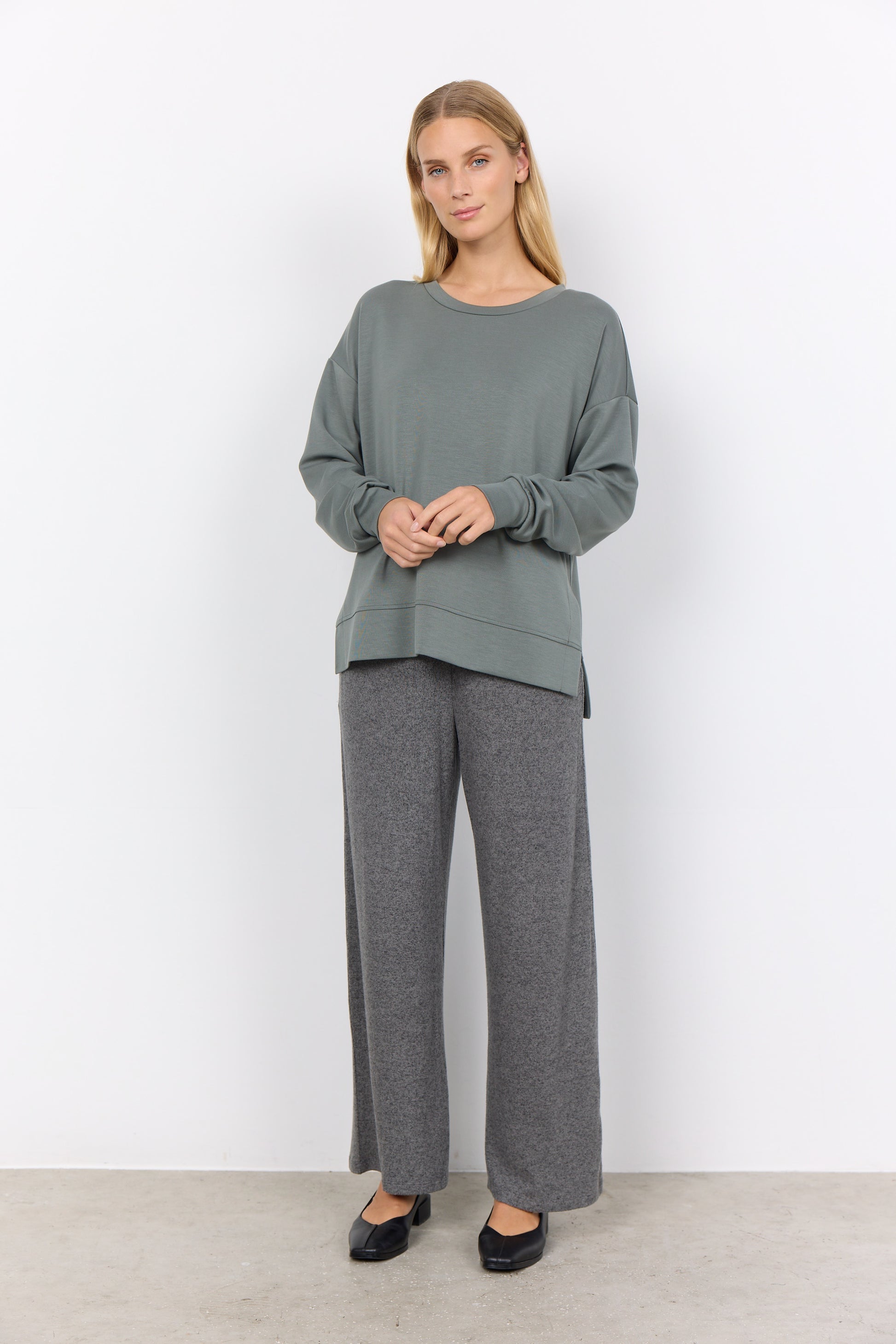 A person wearing a loose-fit Banu 164 Sweater by Soya Concepts in gray stands against a plain white background, paired with pants.