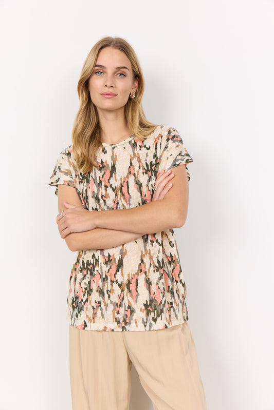 A person with long blonde hair stands against a white background, wearing an abstract print Aretha 38 T-Shirt by Soya Concepts and beige pants, arms crossed.