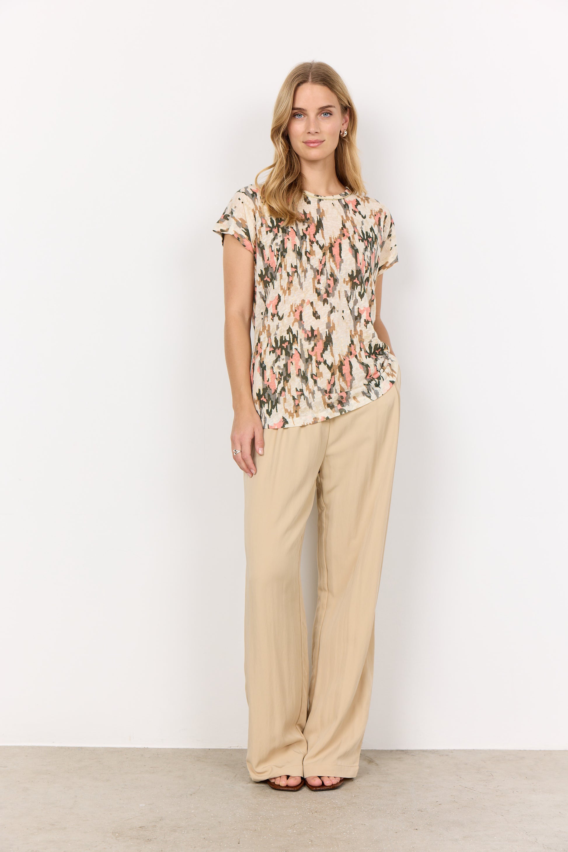 A person with long blonde hair stands against a white background, wearing an abstract print Aretha 38 T-Shirt by Soya Concepts and beige pants, arms crossed.