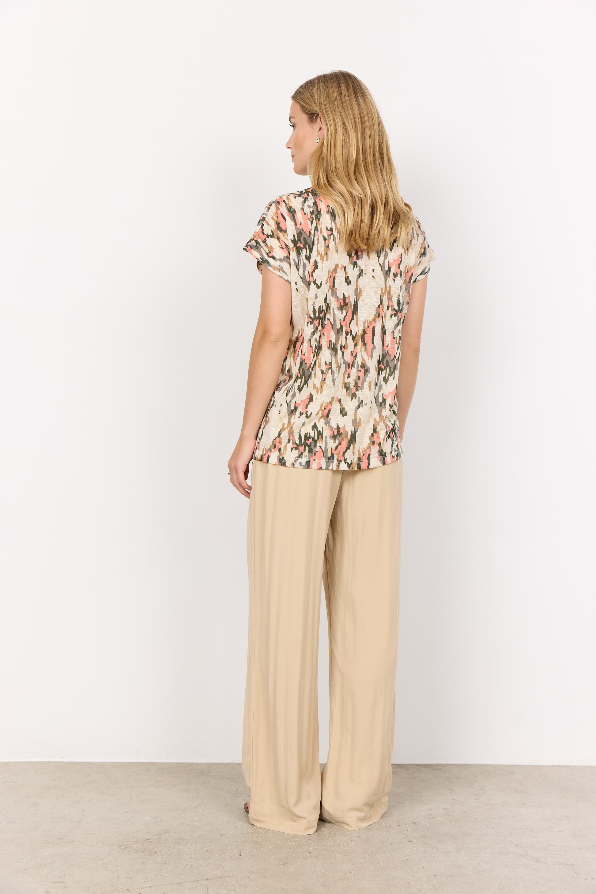 A person with long blonde hair stands against a white background, wearing an abstract print Aretha 38 T-Shirt by Soya Concepts and beige pants, arms crossed.