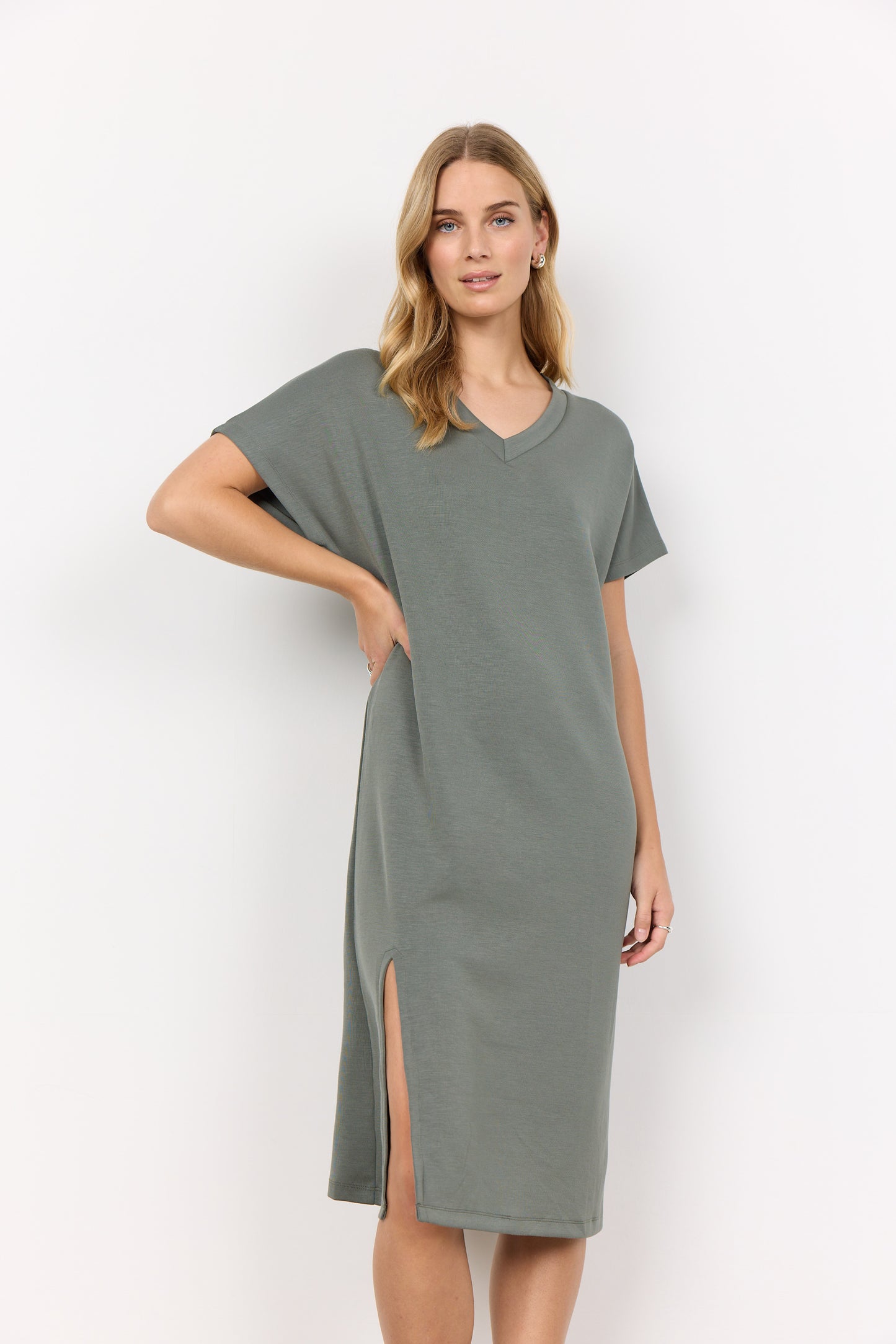 A person stands against a plain white background, wearing the Soya Concepts Banu 218 V-Neck Dress in moss green. This knee-length piece has short sleeves, a relaxed fit, an elegant side slit, and is made from an ultra-soft blend for comfort and style.