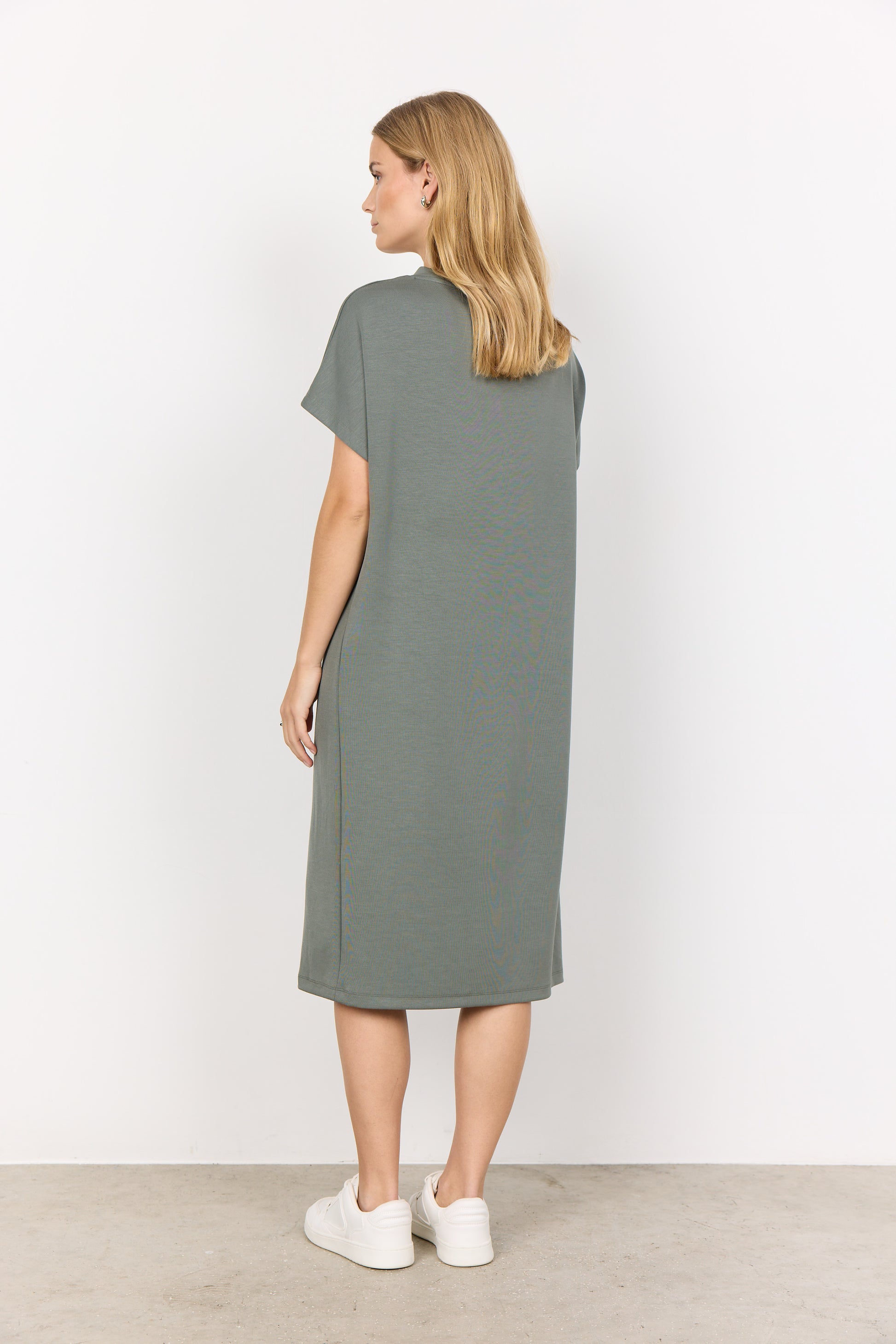 A person stands against a plain white background, wearing the Soya Concepts Banu 218 V-Neck Dress in moss green. This knee-length piece has short sleeves, a relaxed fit, an elegant side slit, and is made from an ultra-soft blend for comfort and style.