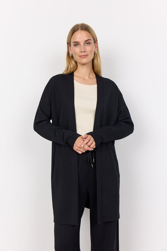 A person with long hair, wearing the Soya Concepts BANU 221 Cardigan in black over a light shirt, stands against a white background. They are looking forward with clasped hands, highlighting the soft fabric's gentle drape and relaxed fit.