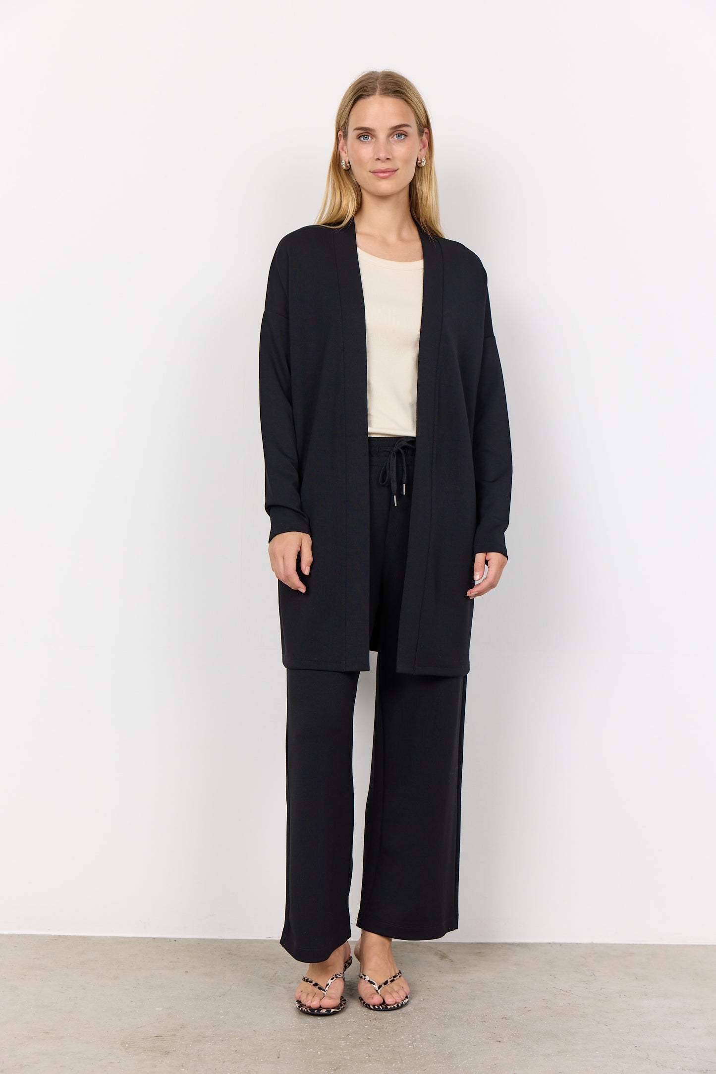 A person with long hair, wearing the Soya Concepts BANU 221 Cardigan in black over a light shirt, stands against a white background. They are looking forward with clasped hands, highlighting the soft fabric's gentle drape and relaxed fit.