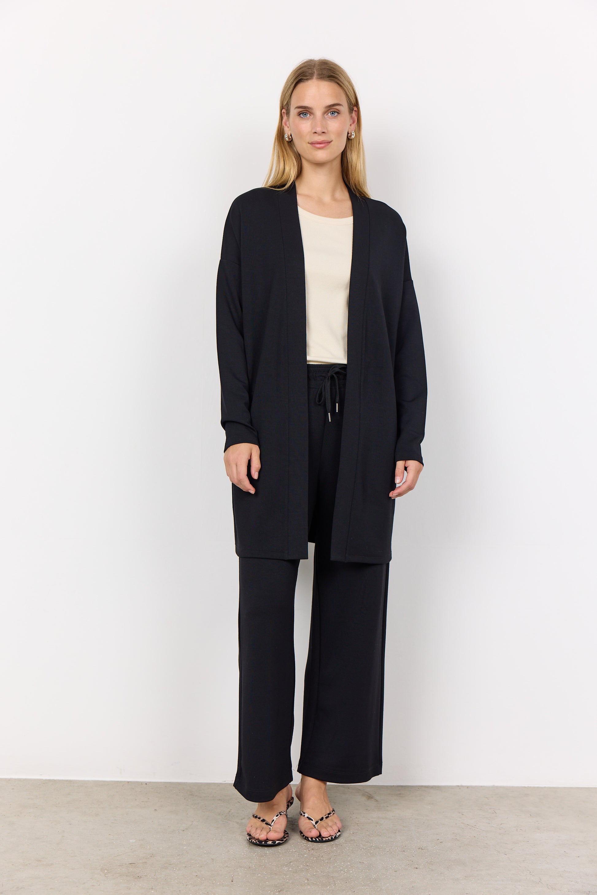 A person with long hair, wearing the Soya Concepts BANU 221 Cardigan in black over a light shirt, stands against a white background. They are looking forward with clasped hands, highlighting the soft fabric's gentle drape and relaxed fit.