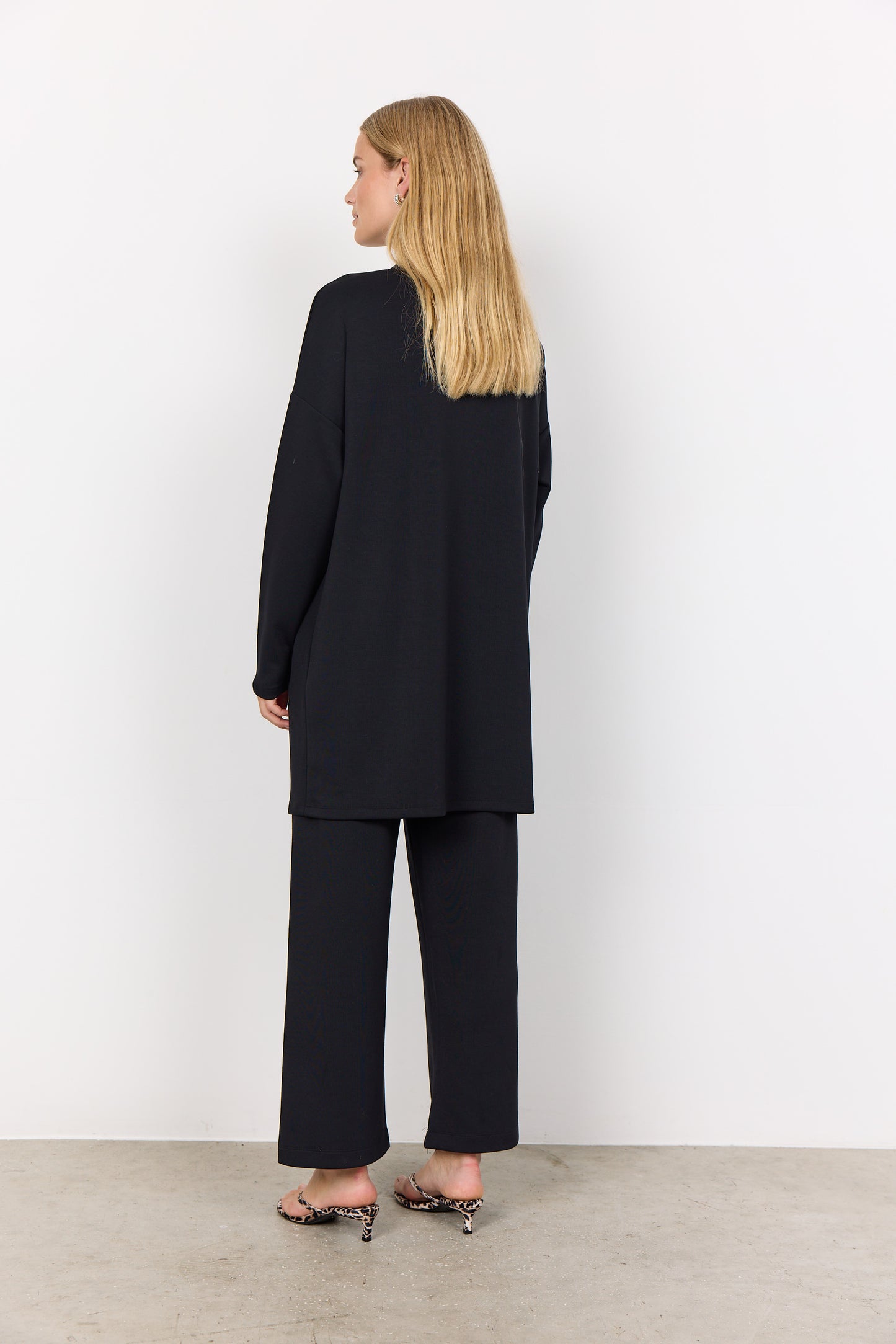 A person with long hair, wearing the Soya Concepts BANU 221 Cardigan in black over a light shirt, stands against a white background. They are looking forward with clasped hands, highlighting the soft fabric's gentle drape and relaxed fit.