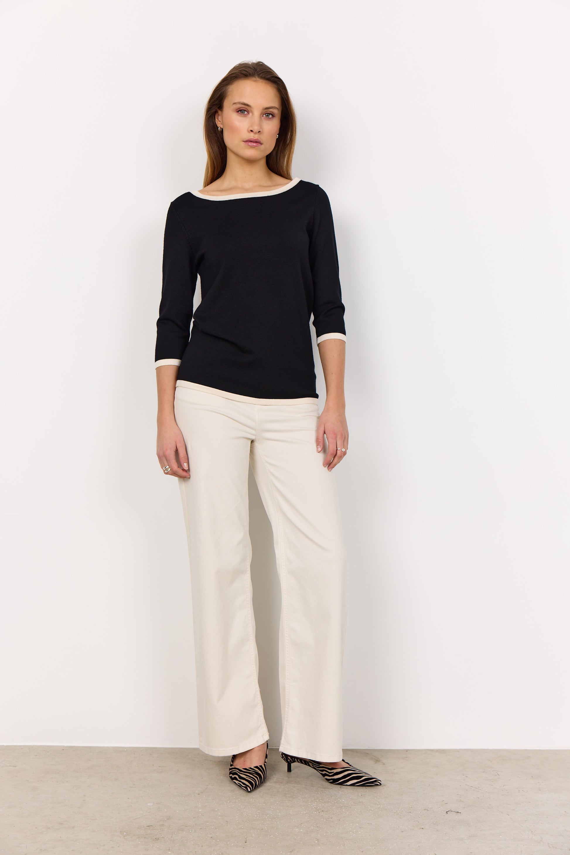 A person models the Soya Concepts Dollie 782 Sweater, featuring a sleek pullover design with a soft knit texture, paired with white pants against a plain white background.