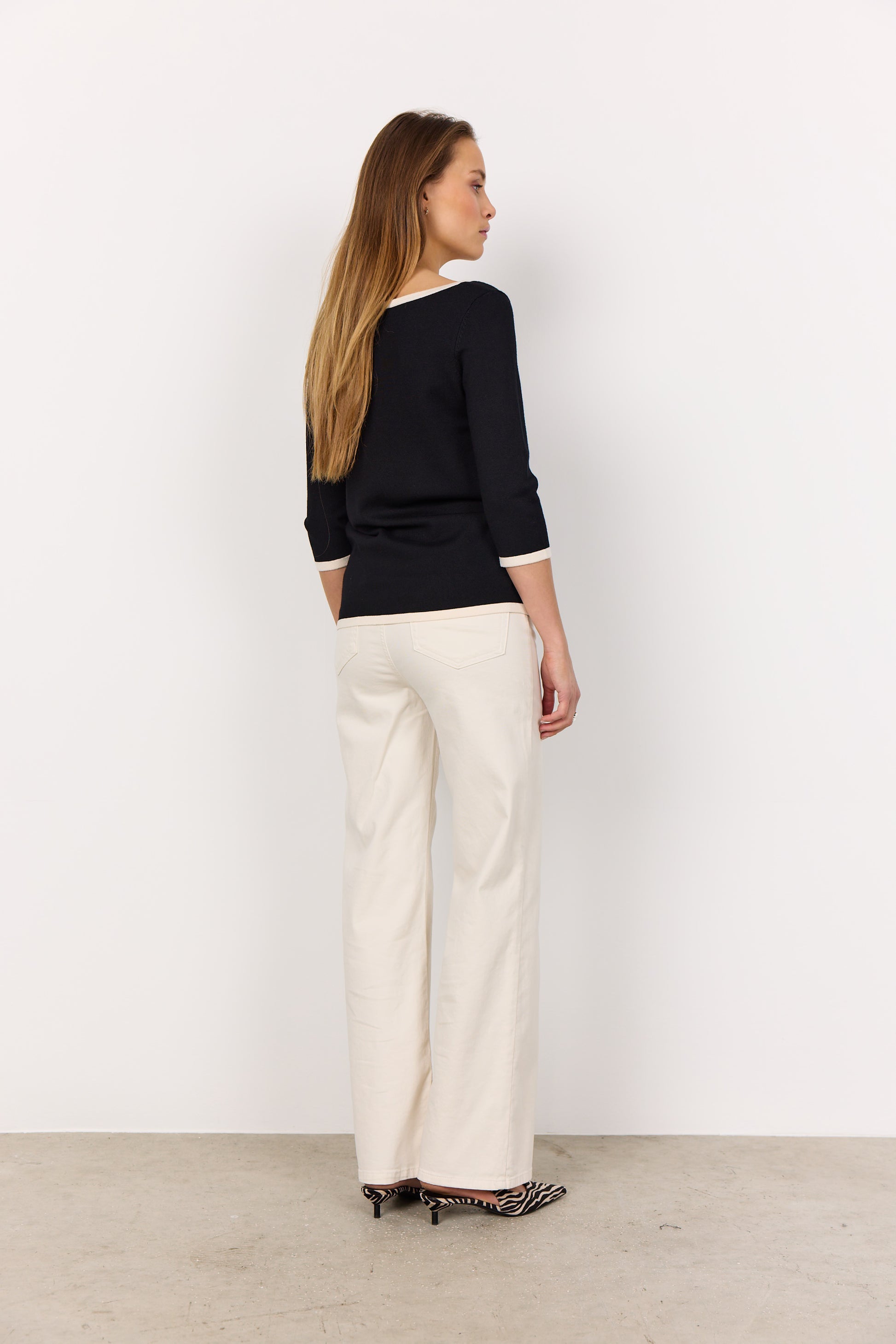 A person models the Soya Concepts Dollie 782 Sweater, featuring a sleek pullover design with a soft knit texture, paired with white pants against a plain white background.