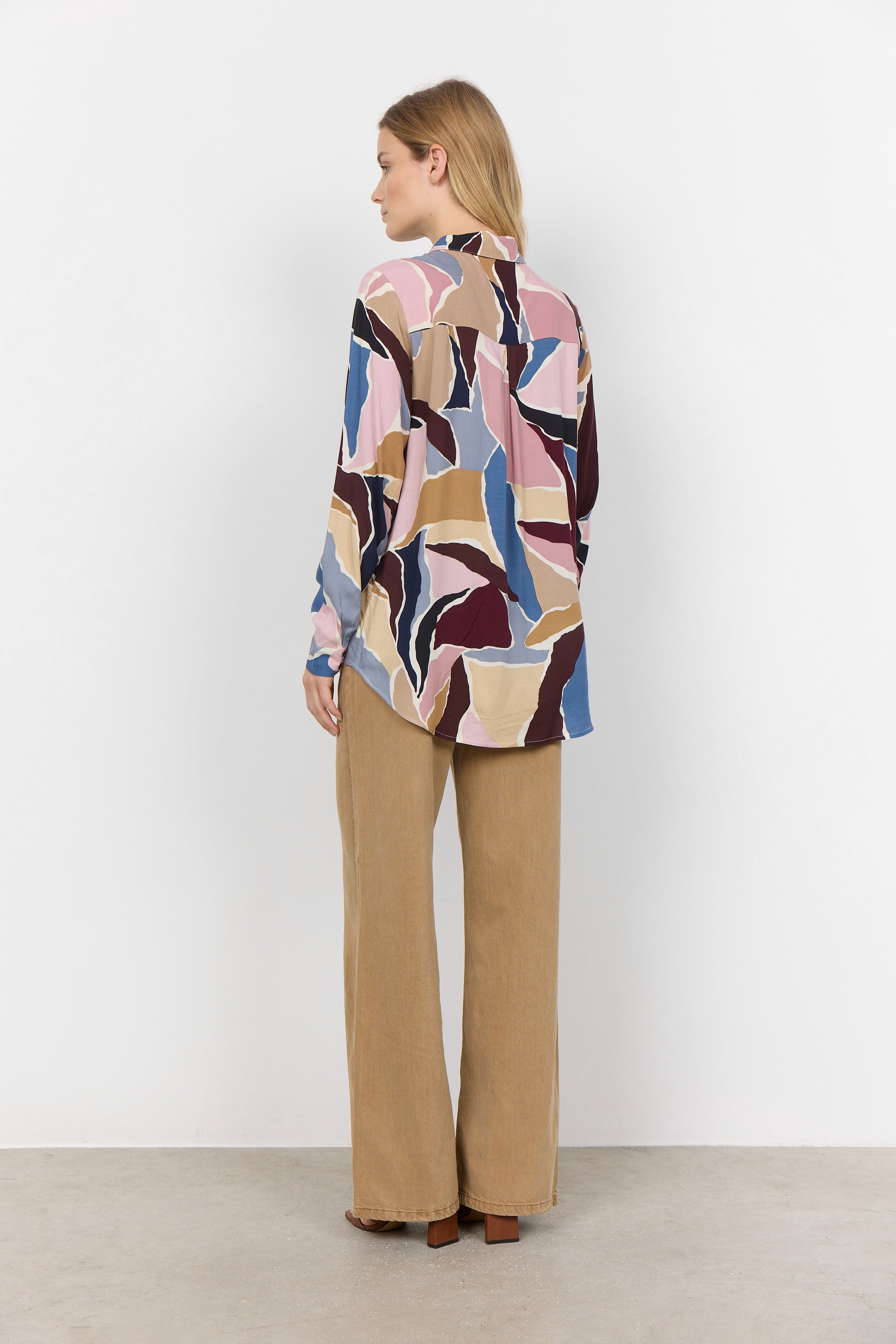 A person stands against a plain background, wearing the ILANA 2 Blouse from Soya Concepts—an abstract-patterned, stylish blouse—paired with beige wide-leg pants and brown open-toed shoes. This versatile wardrobe addition offers a chic and effortless look.