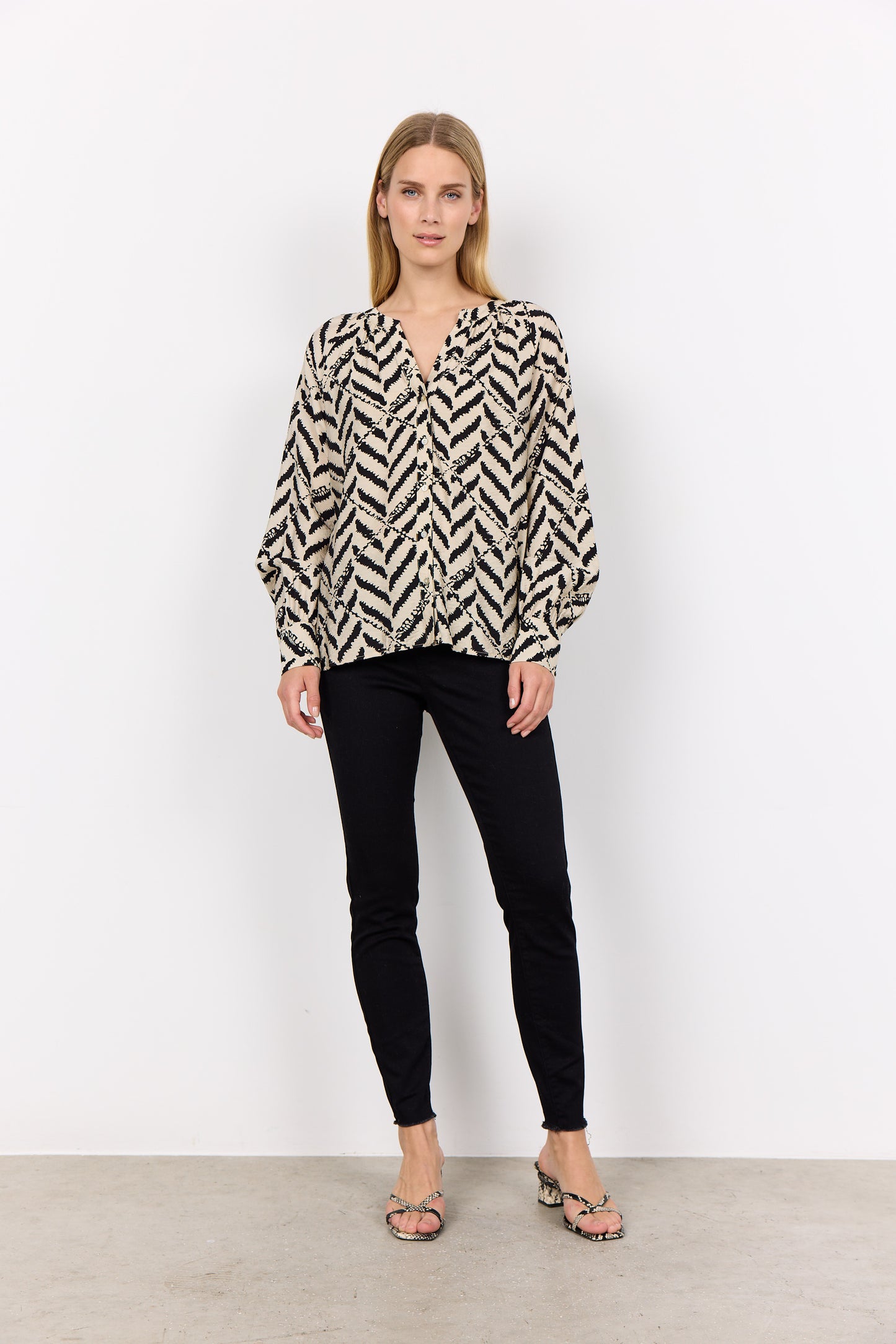 A woman with long hair wears a patterned Inarose 1 Blouse from Soya Concepts and black pants, perfect for an everyday look. She is standing against a plain white background.