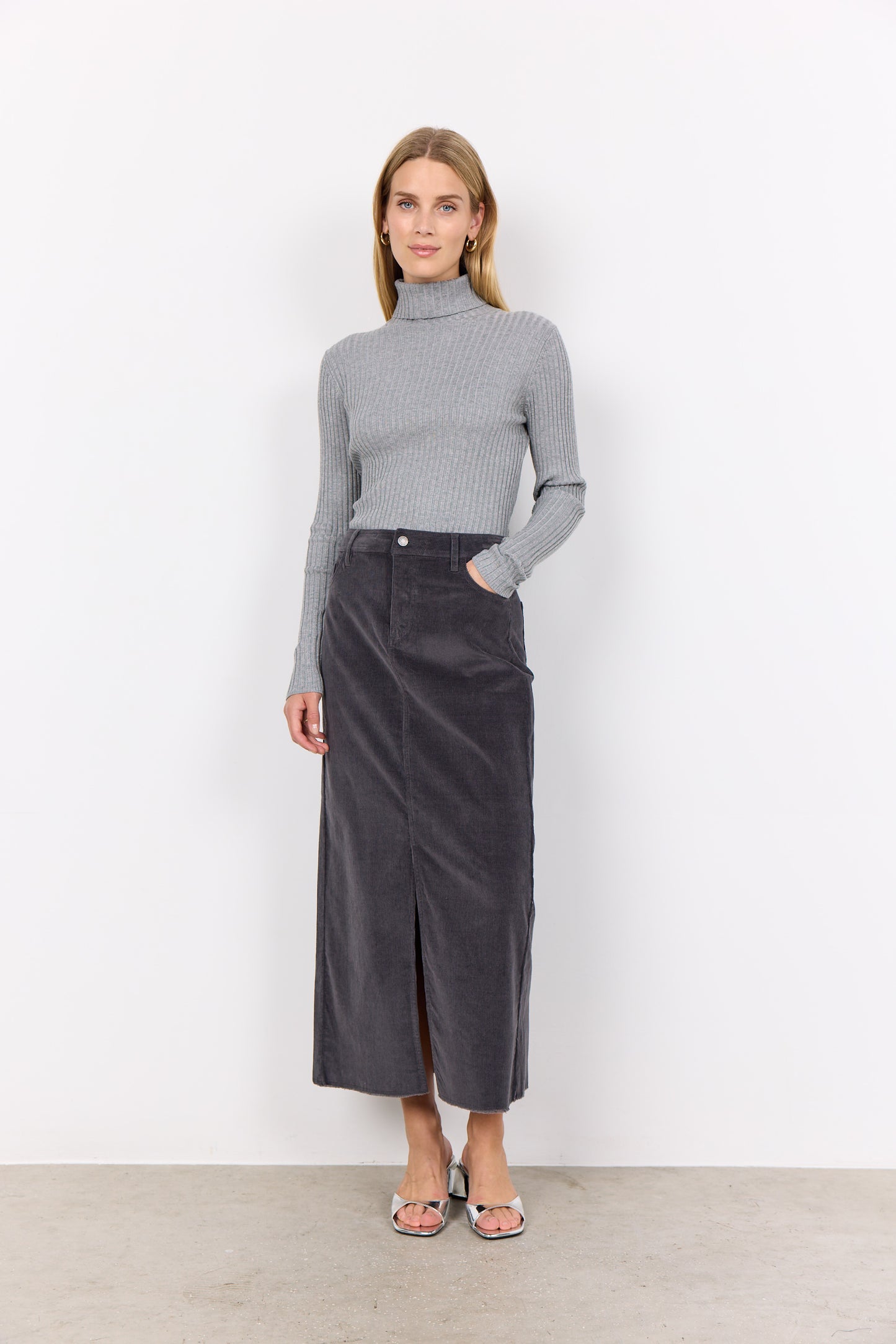 Person wearing a gray corduroy Soya Concepts IVALOU 2 Long Skirt with a front slit and a gray long-sleeve top, standing on a concrete floor in silver open-toe heeled sandals, exuding effortless style.