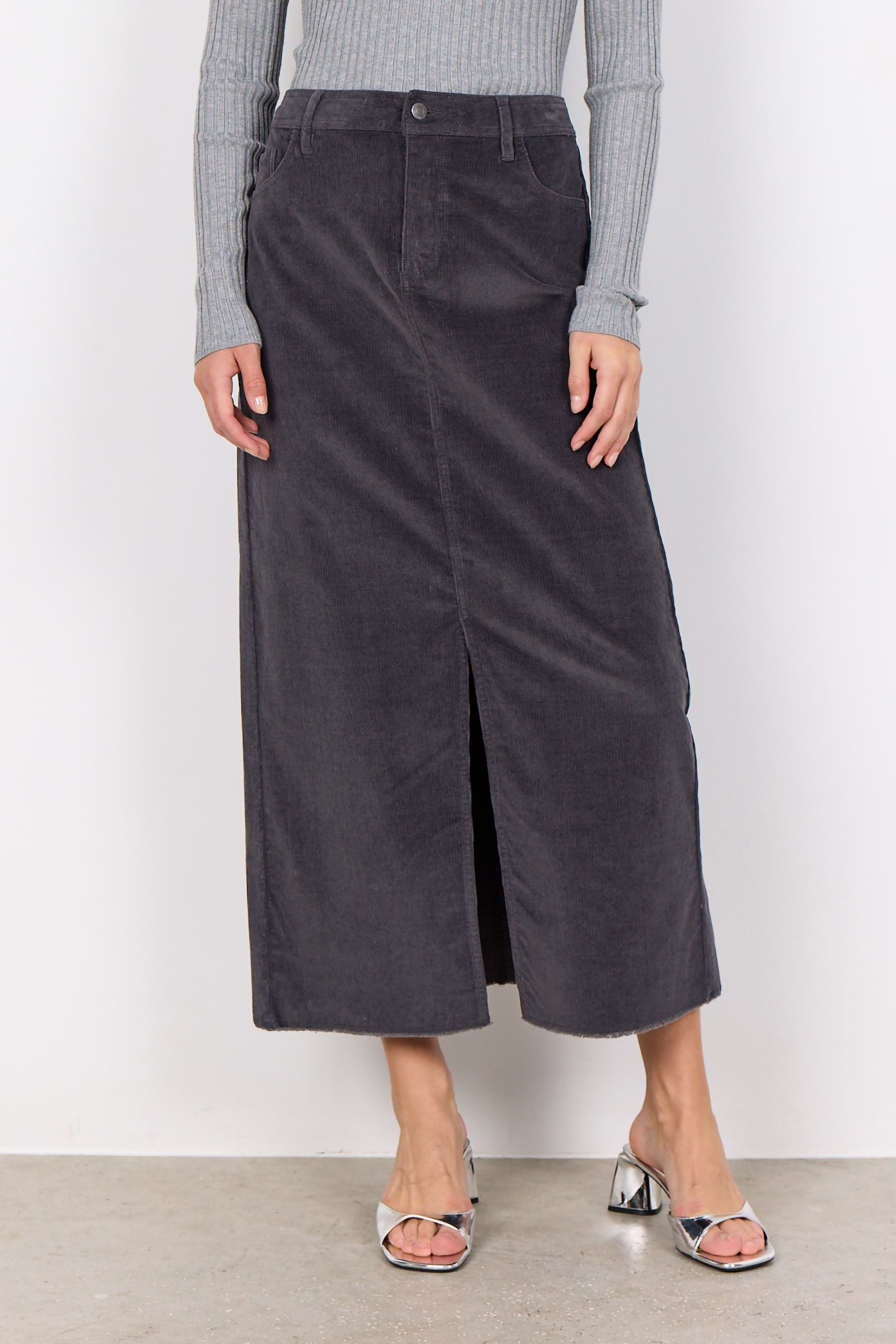 Person wearing a gray corduroy Soya Concepts IVALOU 2 Long Skirt with a front slit and a gray long-sleeve top, standing on a concrete floor in silver open-toe heeled sandals, exuding effortless style.