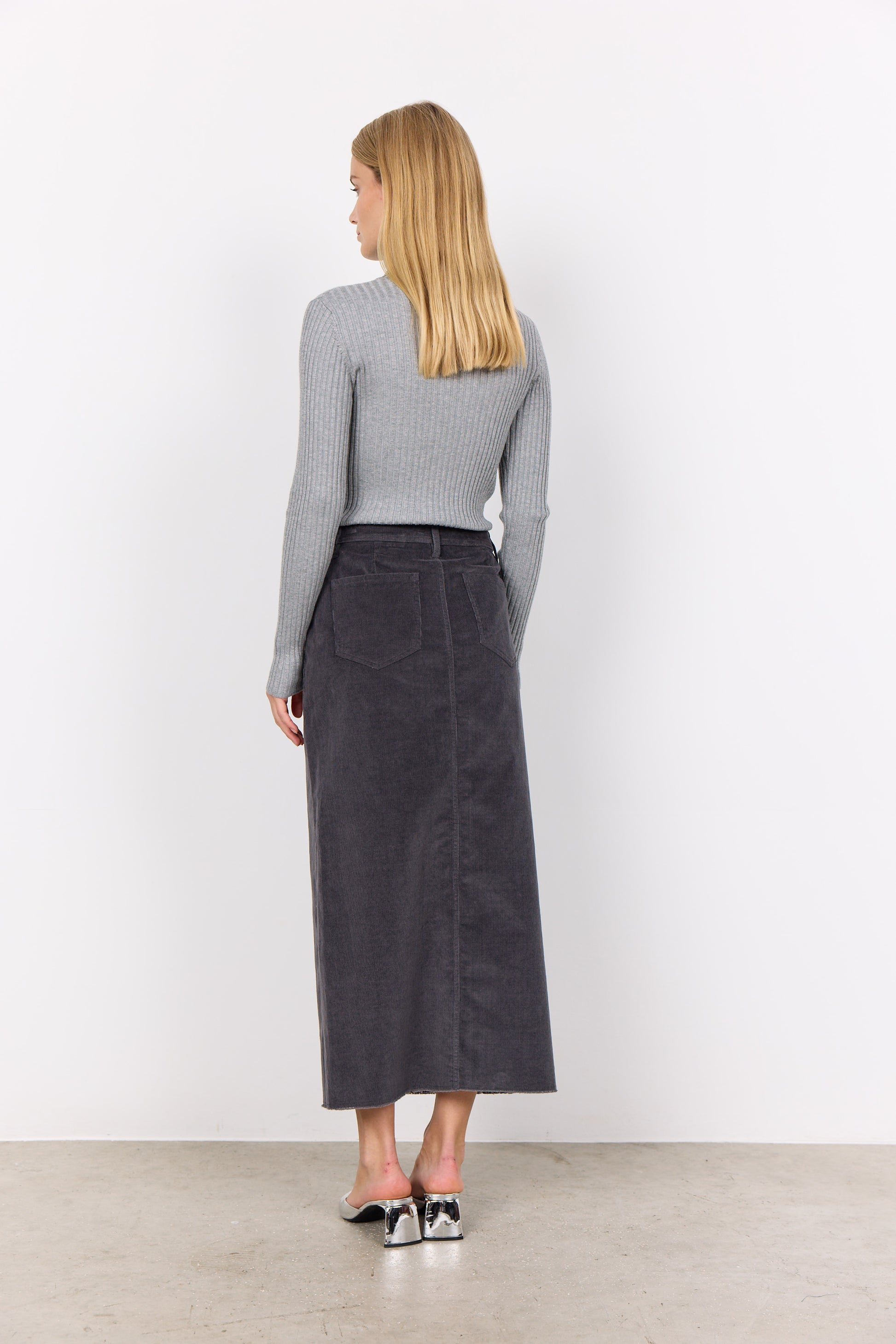 Person wearing a gray corduroy Soya Concepts IVALOU 2 Long Skirt with a front slit and a gray long-sleeve top, standing on a concrete floor in silver open-toe heeled sandals, exuding effortless style.