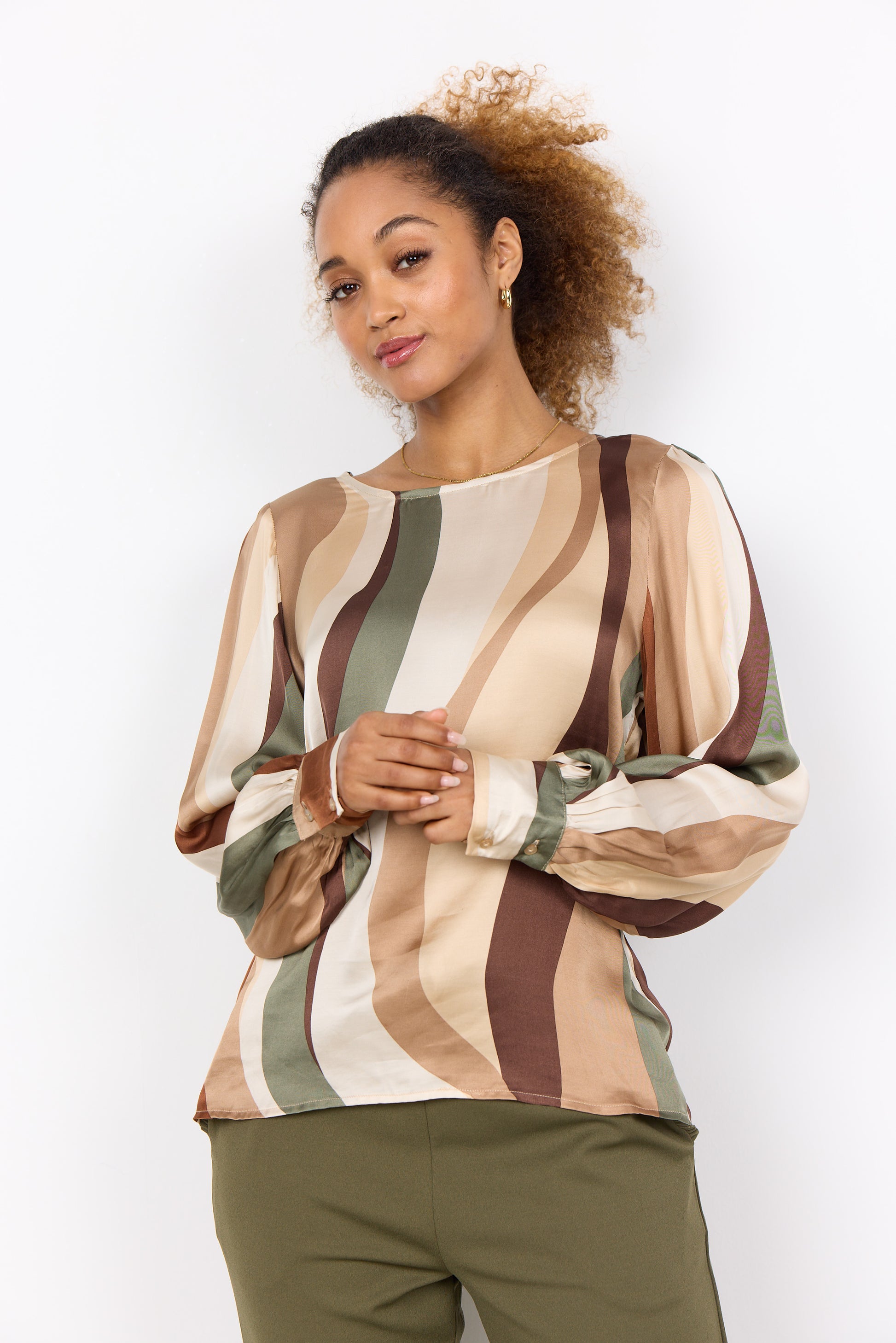 A person with curly hair is wearing a multi-colored, long-sleeved Soya Concepts INES 1 Blouse featuring wavy patterns, paired with olive green pants, creating a trendy look against a plain white background.