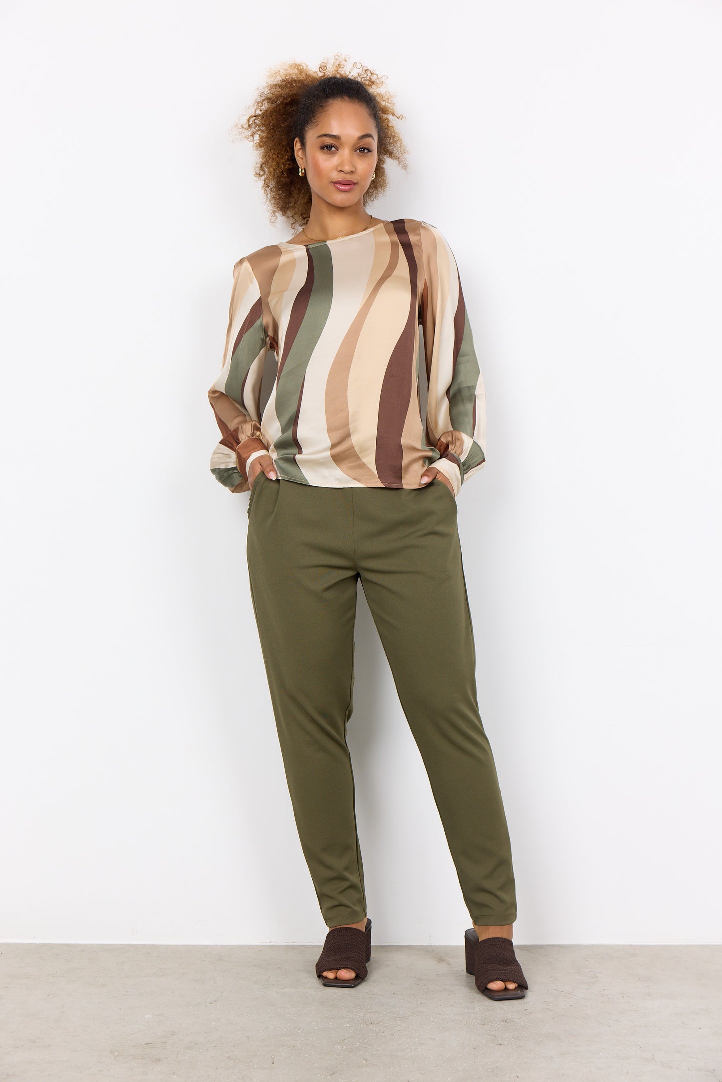 A person with curly hair is wearing a multi-colored, long-sleeved Soya Concepts INES 1 Blouse featuring wavy patterns, paired with olive green pants, creating a trendy look against a plain white background.