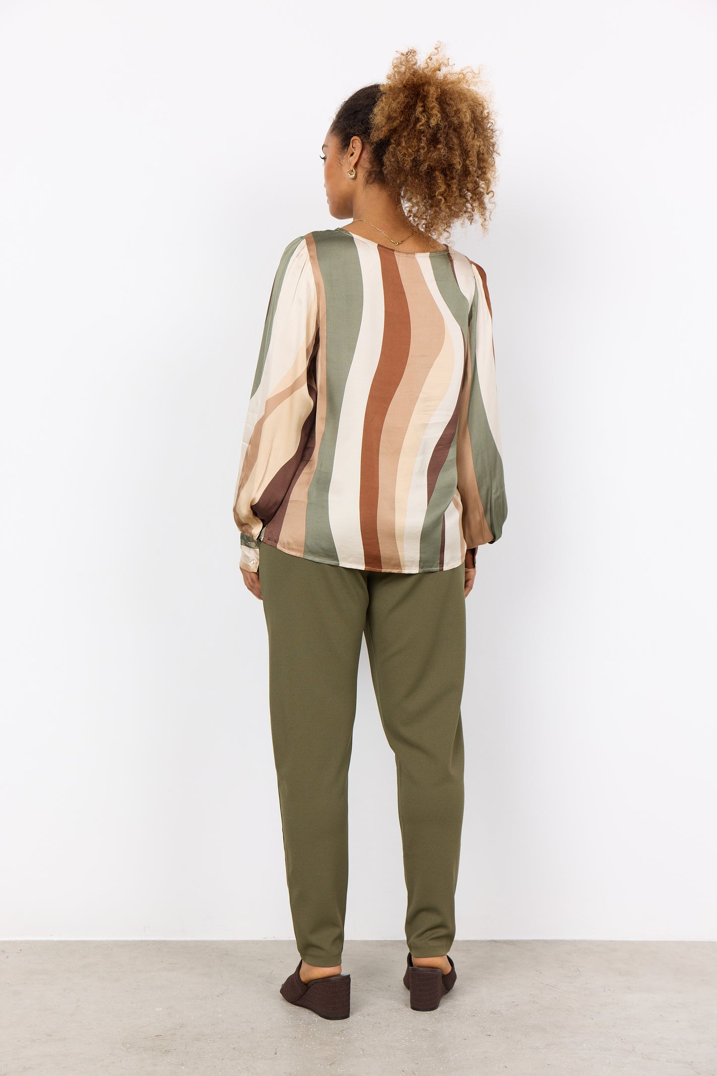 A person with curly hair is wearing a multi-colored, long-sleeved Soya Concepts INES 1 Blouse featuring wavy patterns, paired with olive green pants, creating a trendy look against a plain white background.