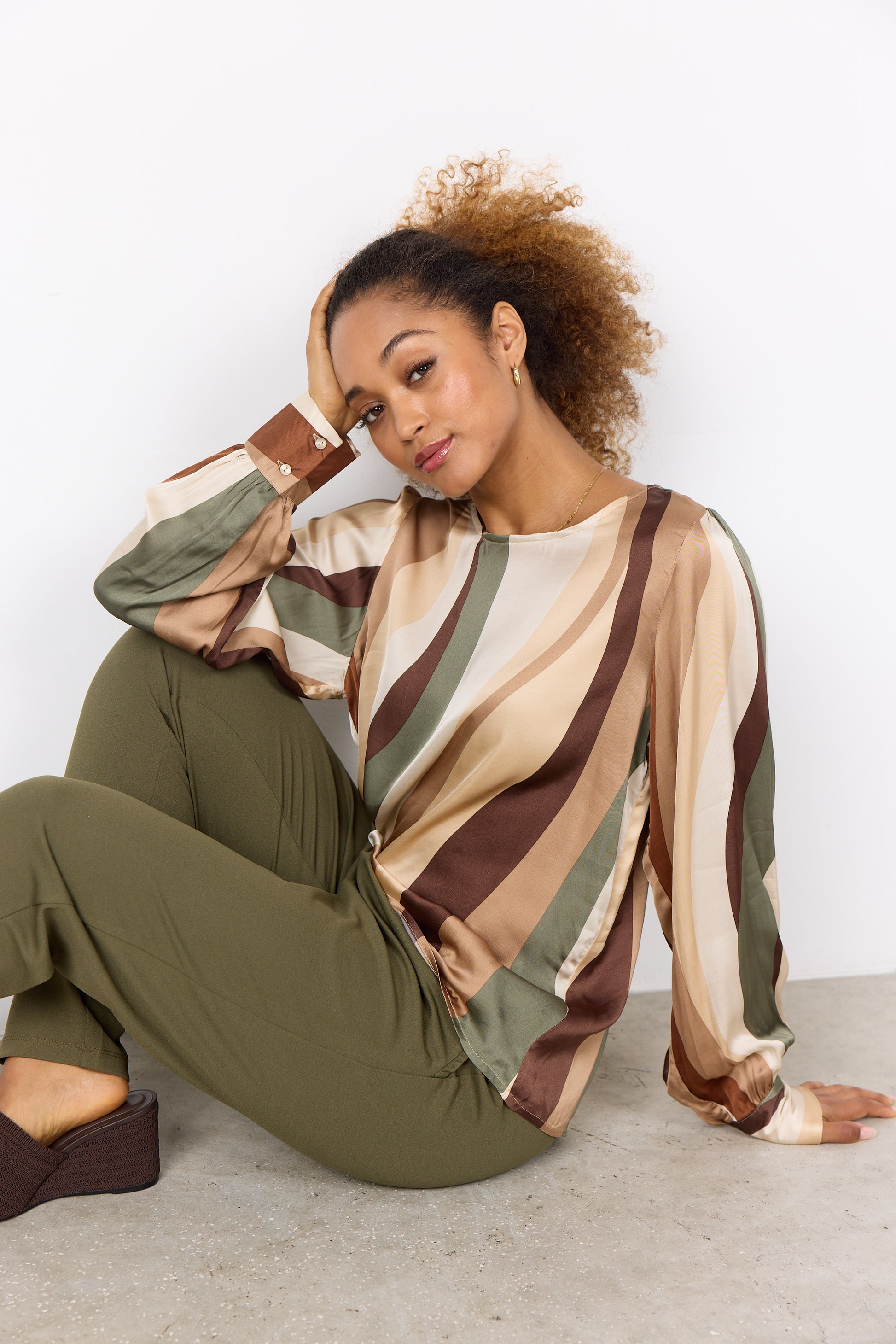 A person with curly hair is wearing a multi-colored, long-sleeved Soya Concepts INES 1 Blouse featuring wavy patterns, paired with olive green pants, creating a trendy look against a plain white background.