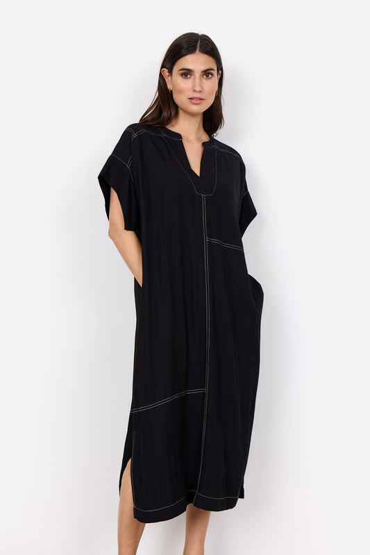 Someone is wearing a Paige 3 Midi Dress by Soya Concepts, which is loose-fitting and black with white stitching and a V-neckline, standing against a white background.