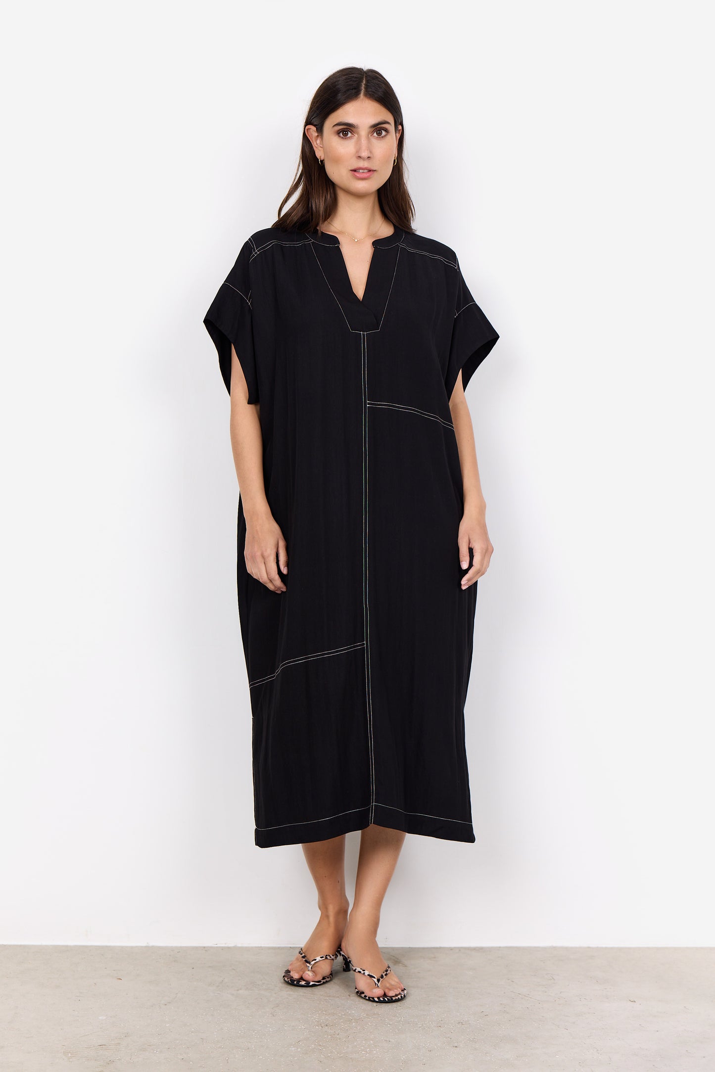 Someone is wearing a Paige 3 Midi Dress by Soya Concepts, which is loose-fitting and black with white stitching and a V-neckline, standing against a white background.