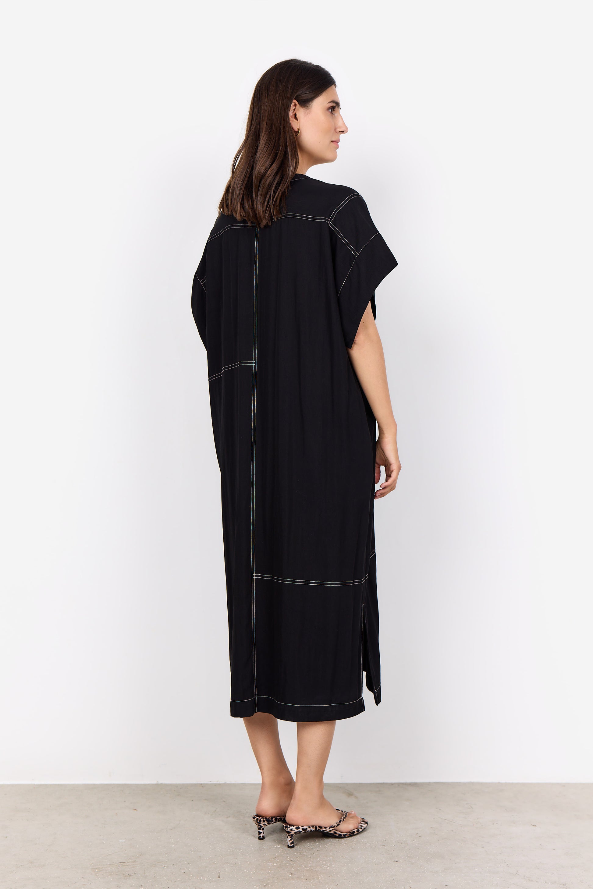 Someone is wearing a Paige 3 Midi Dress by Soya Concepts, which is loose-fitting and black with white stitching and a V-neckline, standing against a white background.