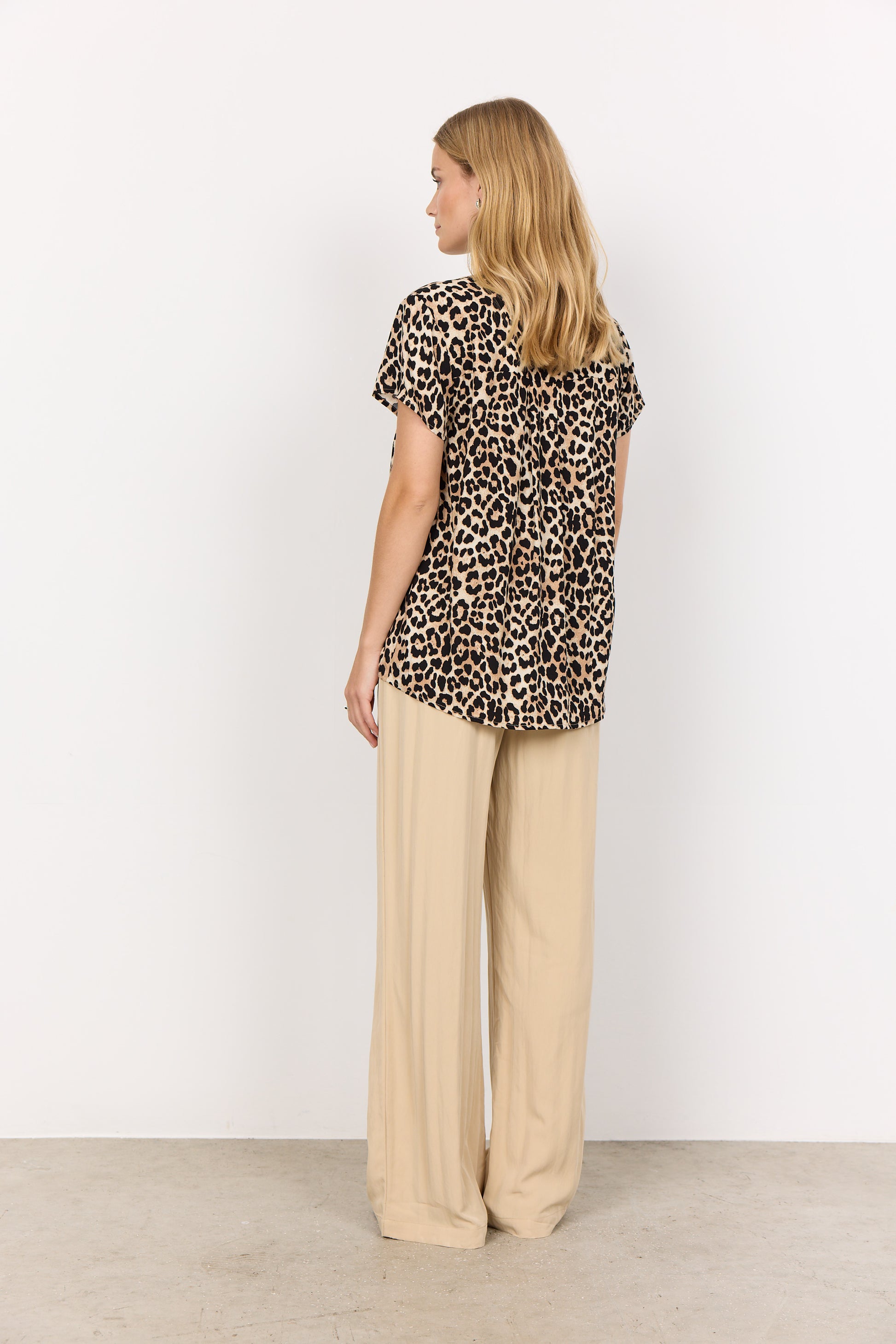 A person confidently stands with folded arms against a white background, wearing a striking leopard print Pietta Blouse by Soya Concepts and beige pants.