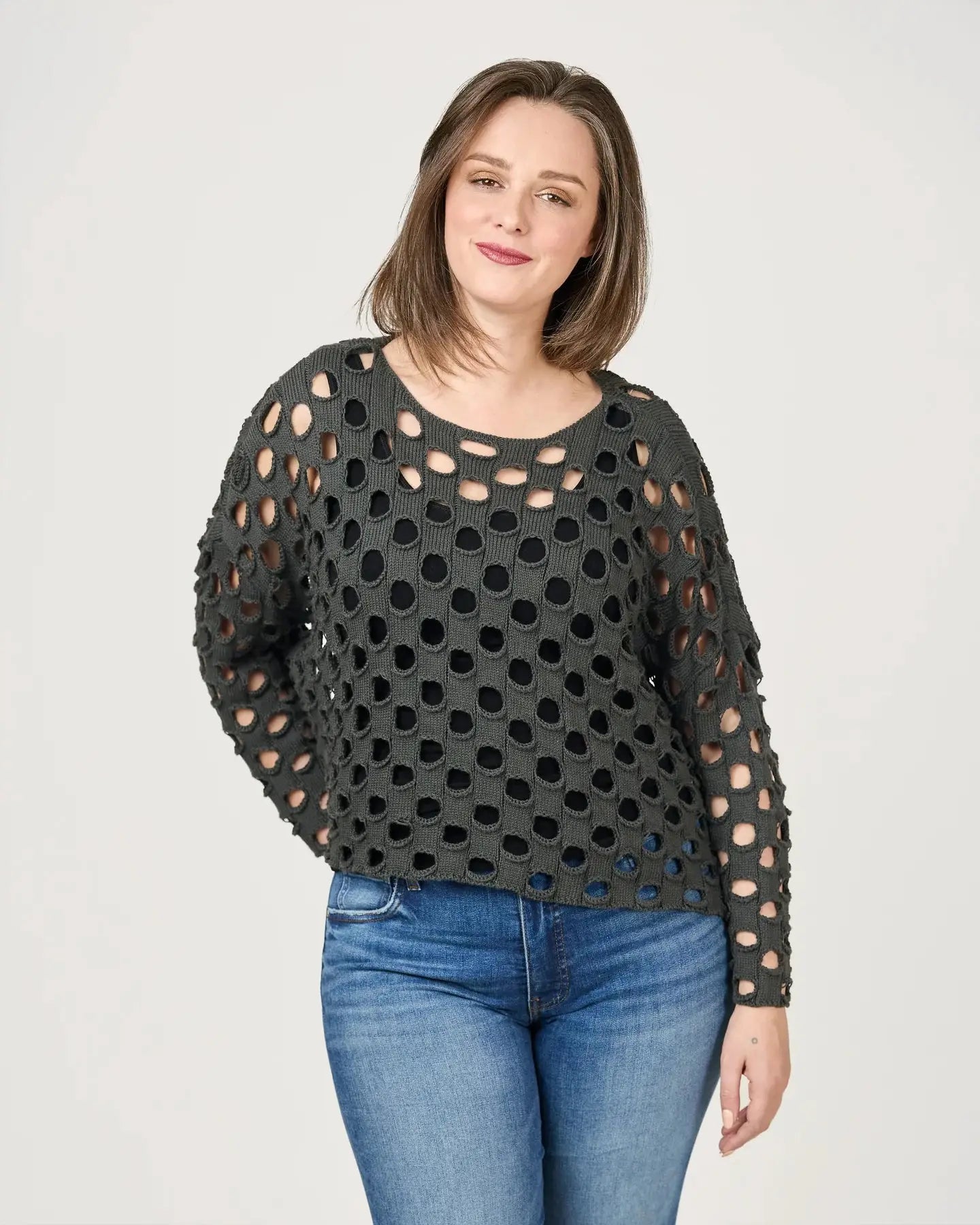 A person with shoulder-length hair, dressed in a chic black open-knit Lula Pullover by Shannon Passero and blue jeans, stands against a plain background.