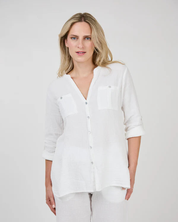 A person with long, blonde hair, wearing a ROXIE Woven Top by Shannon Passero made from quality materials stands against a plain background, showcasing a stylish look in their white button-up shirt with long sleeves.