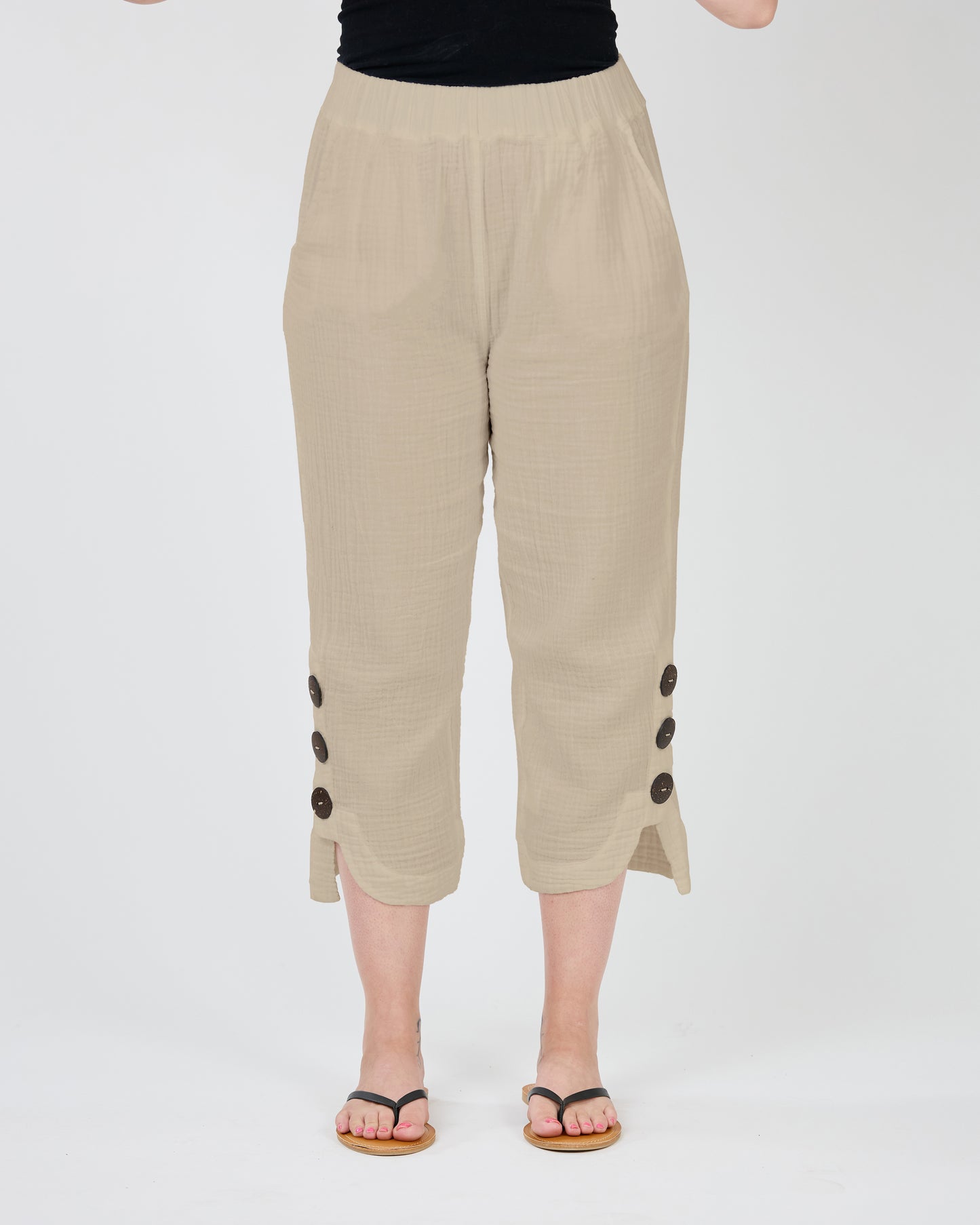 Person standing, showcasing beige Shannon Passero JUNIPER Crop Pant with button details on the sides and wearing black flip-flops, against a white background. These must-have pants are both stylish and functional.