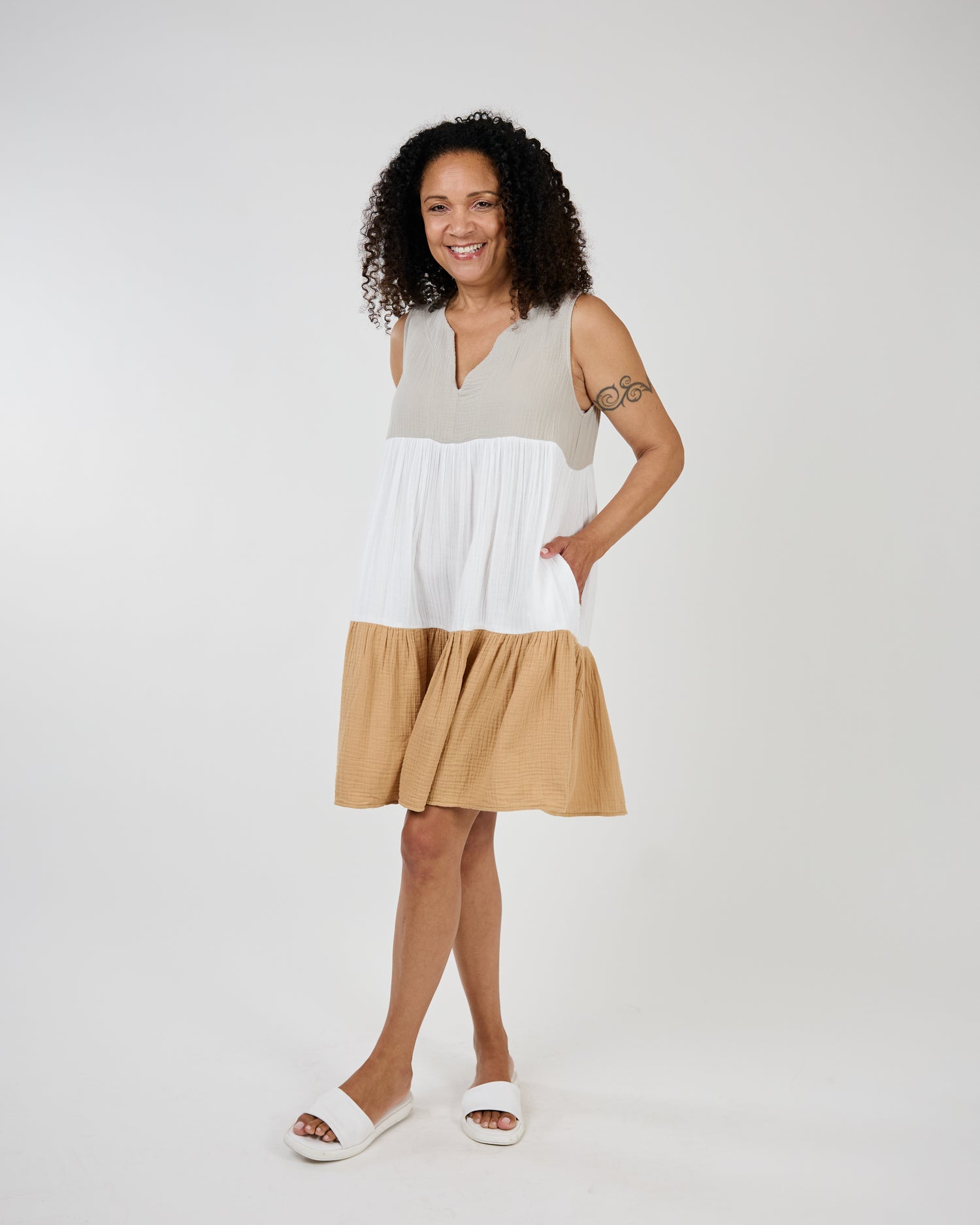 Woman smiling in a sleeveless, Shannon Passero Sandmix Lyric Mid Length Dress and white sandals against a plain background.