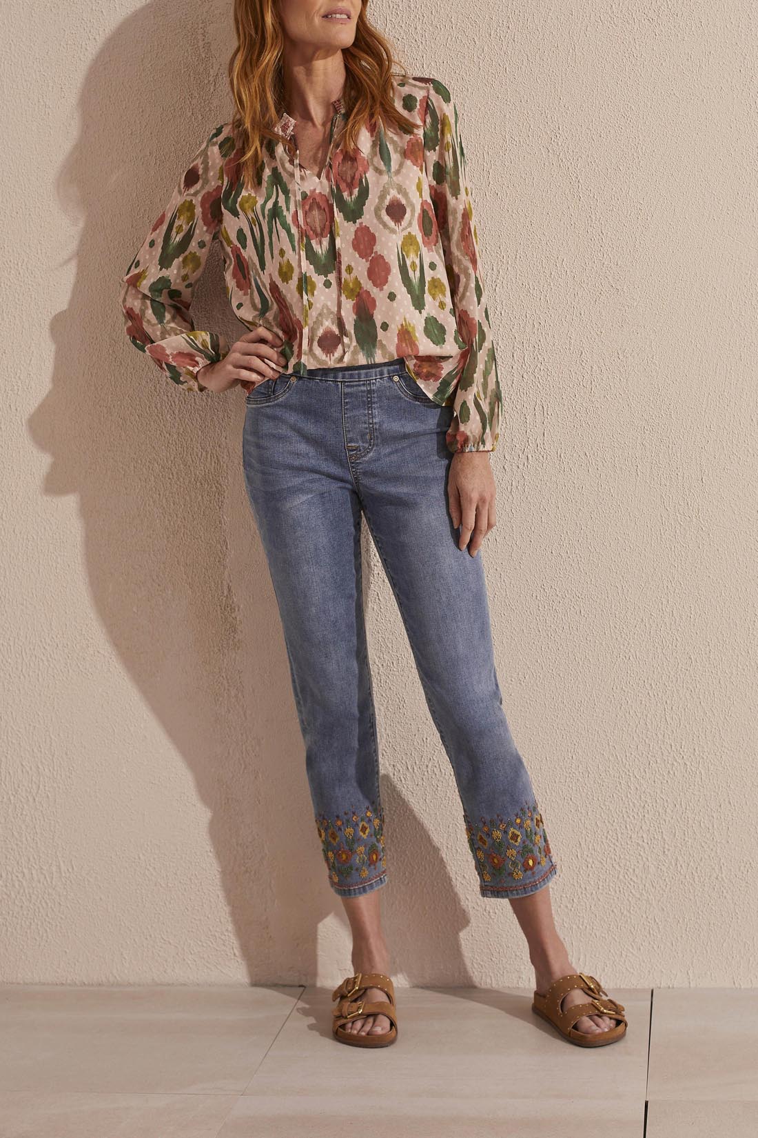 A woman stands wearing a floral print blouse and Tribal's Embroidered Pull On Straight Ankle Jeans with Beading paired with gold sandals.