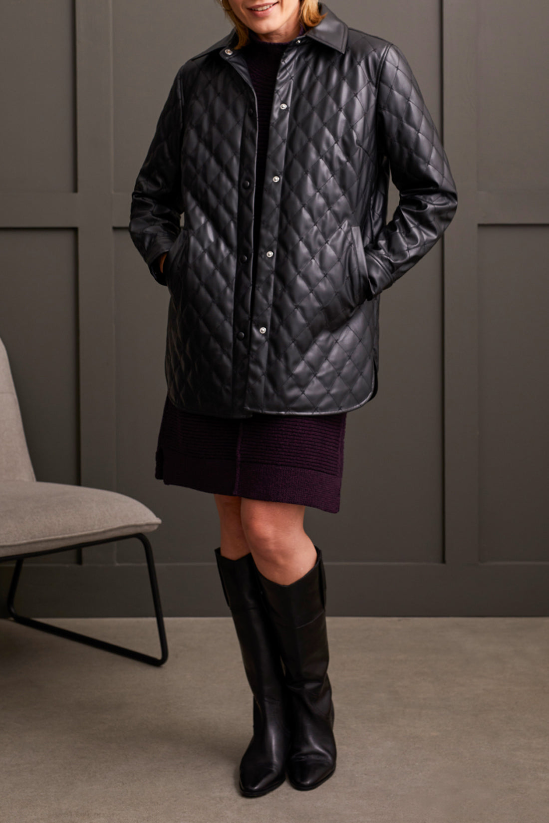 In a room with gray paneled walls, an individual is seen wearing a warm and fashionable black Quilted Snap Front Puffer Jacket from Tribal, paired with knee-high black boots. Made from high-quality materials, the jacket enhances the look alongside the partially visible chair in the background.