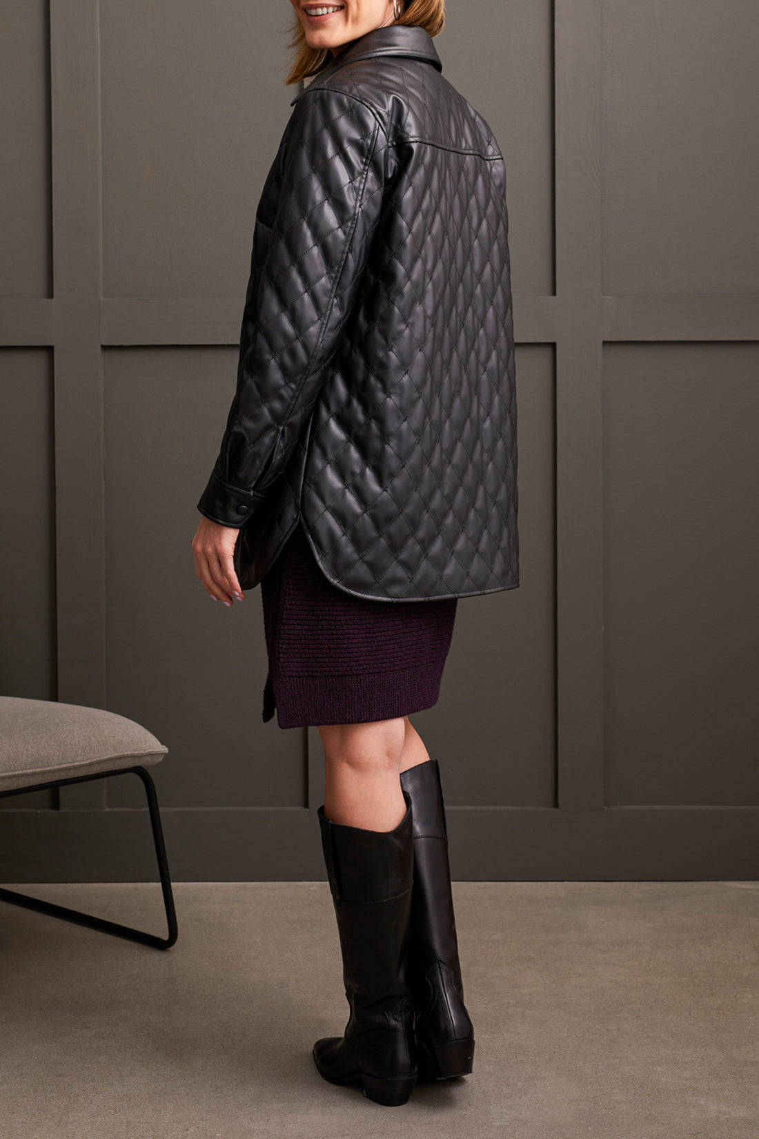 In a room with gray paneled walls, an individual is seen wearing a warm and fashionable black Quilted Snap Front Puffer Jacket from Tribal, paired with knee-high black boots. Made from high-quality materials, the jacket enhances the look alongside the partially visible chair in the background.