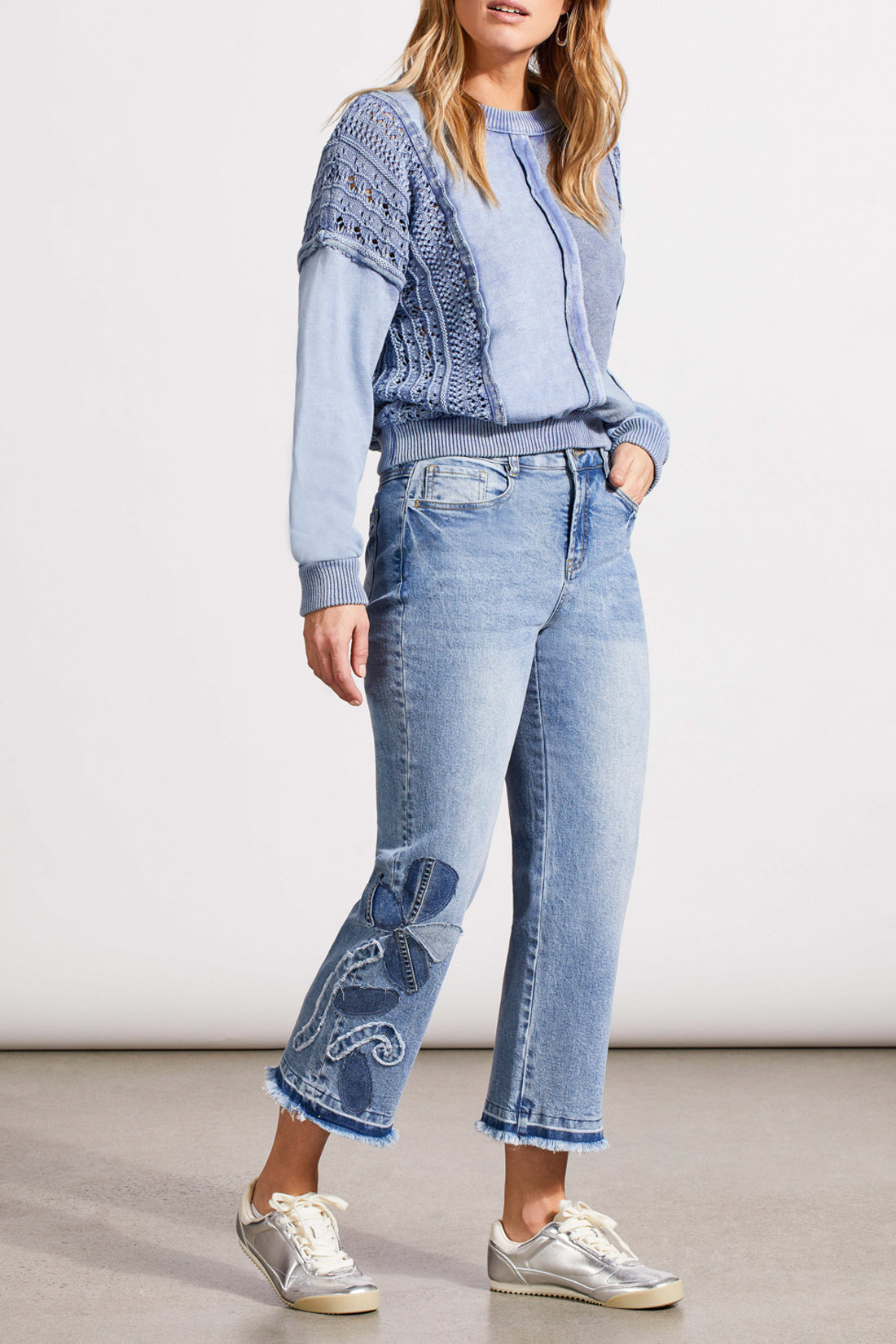 A person is wearing a light blue, textured sweater, Tribal's Sophia Microflare Crop with Denim jeans featuring embroidery near the hem, and silver sneakers. The image captures the outfit from head to lower legs.