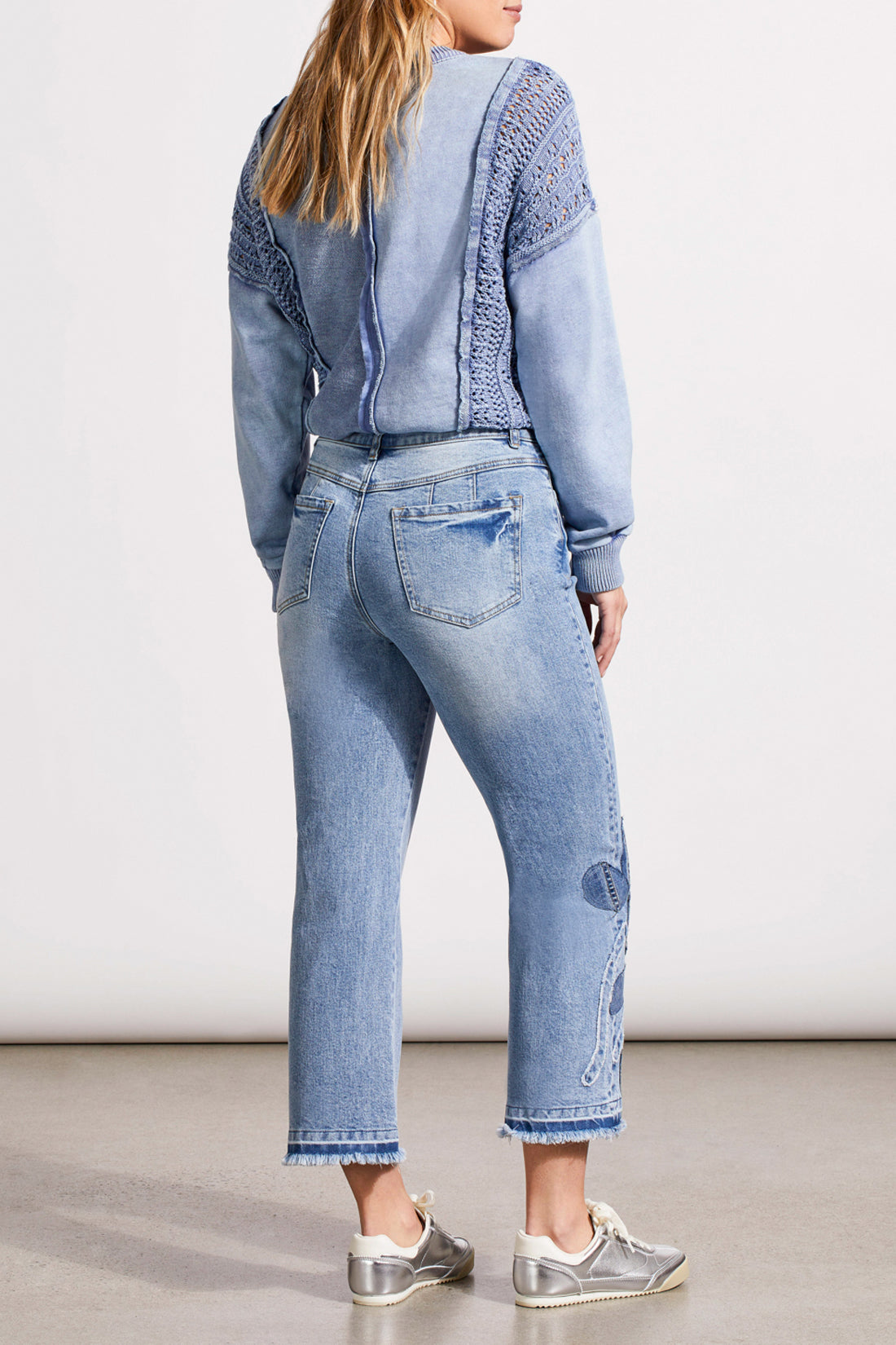 A person is wearing a light blue, textured sweater, Tribal's Sophia Microflare Crop with Denim jeans featuring embroidery near the hem, and silver sneakers. The image captures the outfit from head to lower legs.