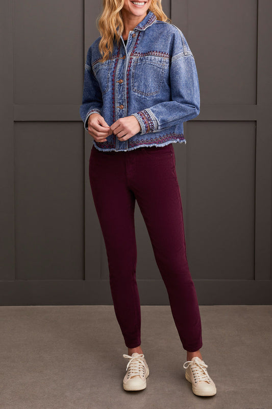 A woman is wearing the Tribal Crop Denim Shacket with Embroidery, featuring a frayed hem, paired with burgundy pants and beige sneakers, standing in front of a dark paneled wall.