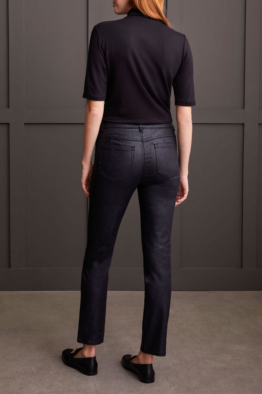 A person stands against a gray wall wearing a black turtleneck, Sophia Hugging Caviar Glitz Slim Jeans by Tribal, and black loafers, exuding playful elegance. Their right hand is casually tucked in their pocket.