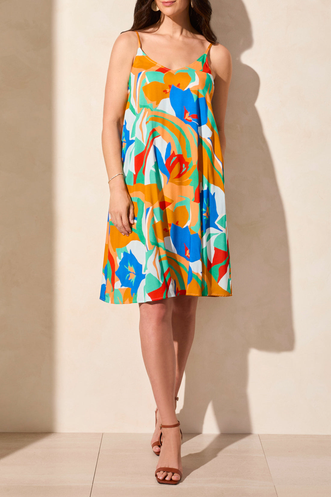 A woman models the Tribal UNLINED DRESS W/ POCKETS against a neutral background, showcasing its brightly colored floral design. Her neck is not visible in the image.