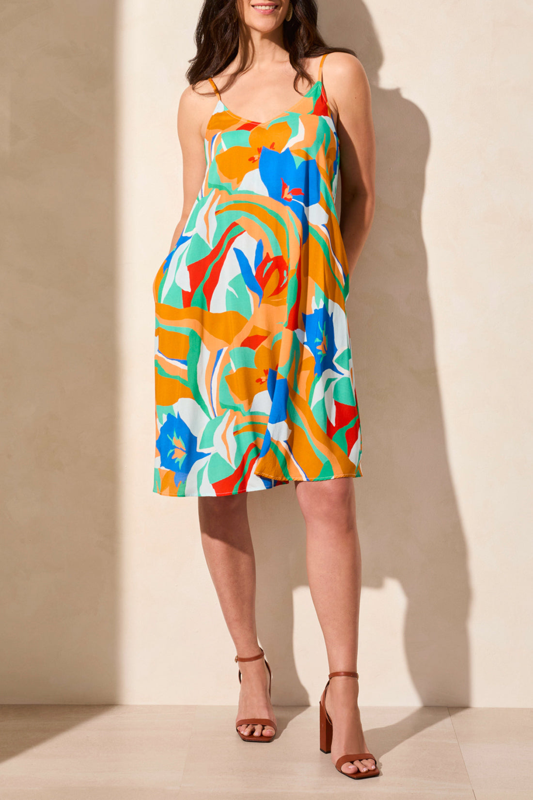 A woman models the Tribal UNLINED DRESS W/ POCKETS against a neutral background, showcasing its brightly colored floral design. Her neck is not visible in the image.